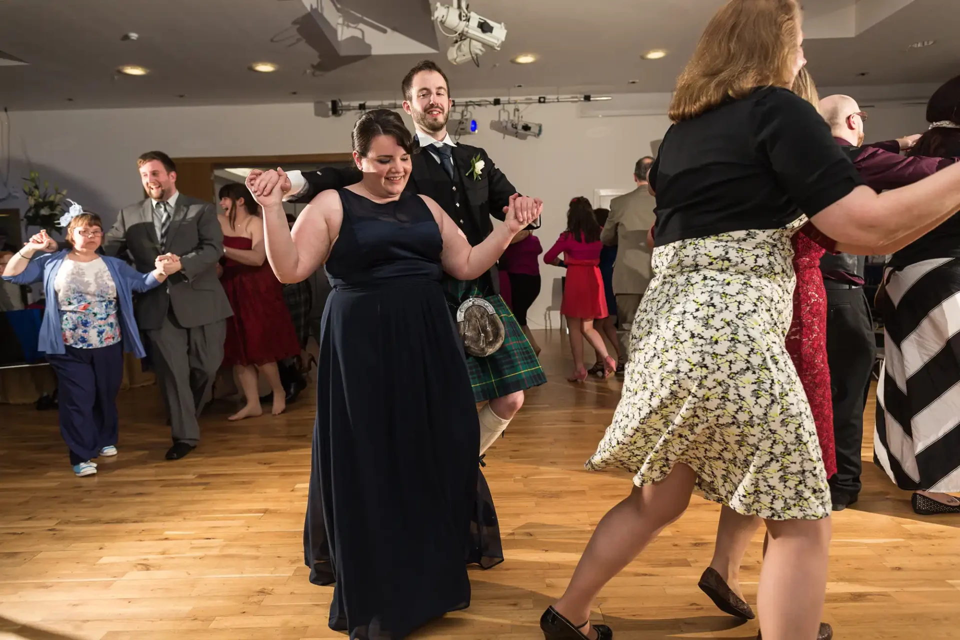 Guests enjoy dancing at a lively indoor wedding reception, with a man in a kilt and woman in a blue dress smiling and holding hands in the foreground.