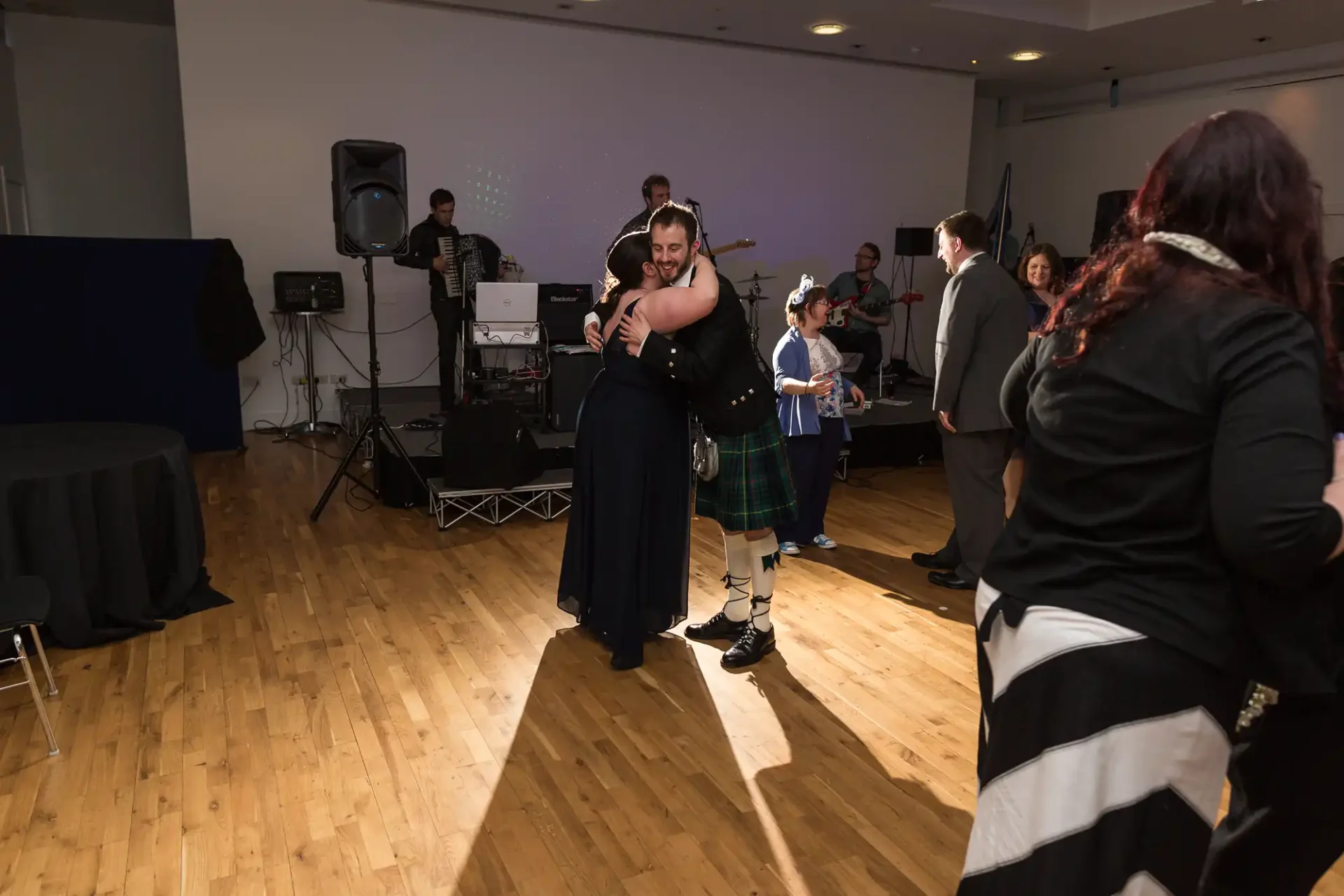 A couple dancing joyfully at a lively indoor event while a band performs in the background and others watch.
