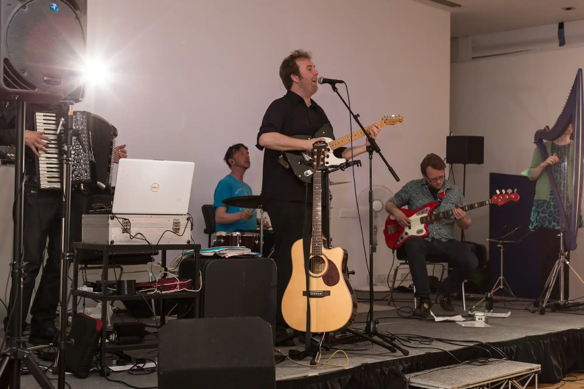 A band performs at an indoor event, featuring a vocalist, guitarist, bassist, keyboardist with a laptop, and an accordion player.
