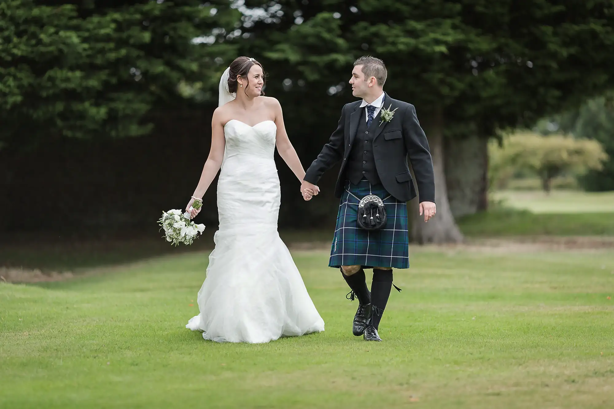 Bride in a white dress and groom in a kilt holding hands and walking through a grassy field, smiling at each other.