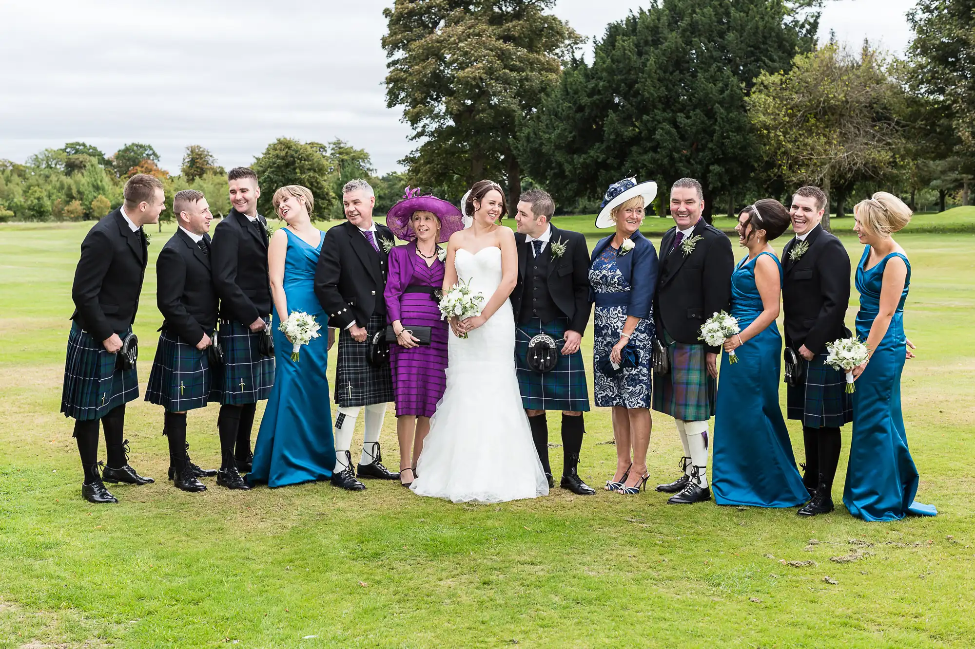 A wedding group in formal attire with kilts and dresses, smiling outdoors on a grassy field, with some guests looking at each other.