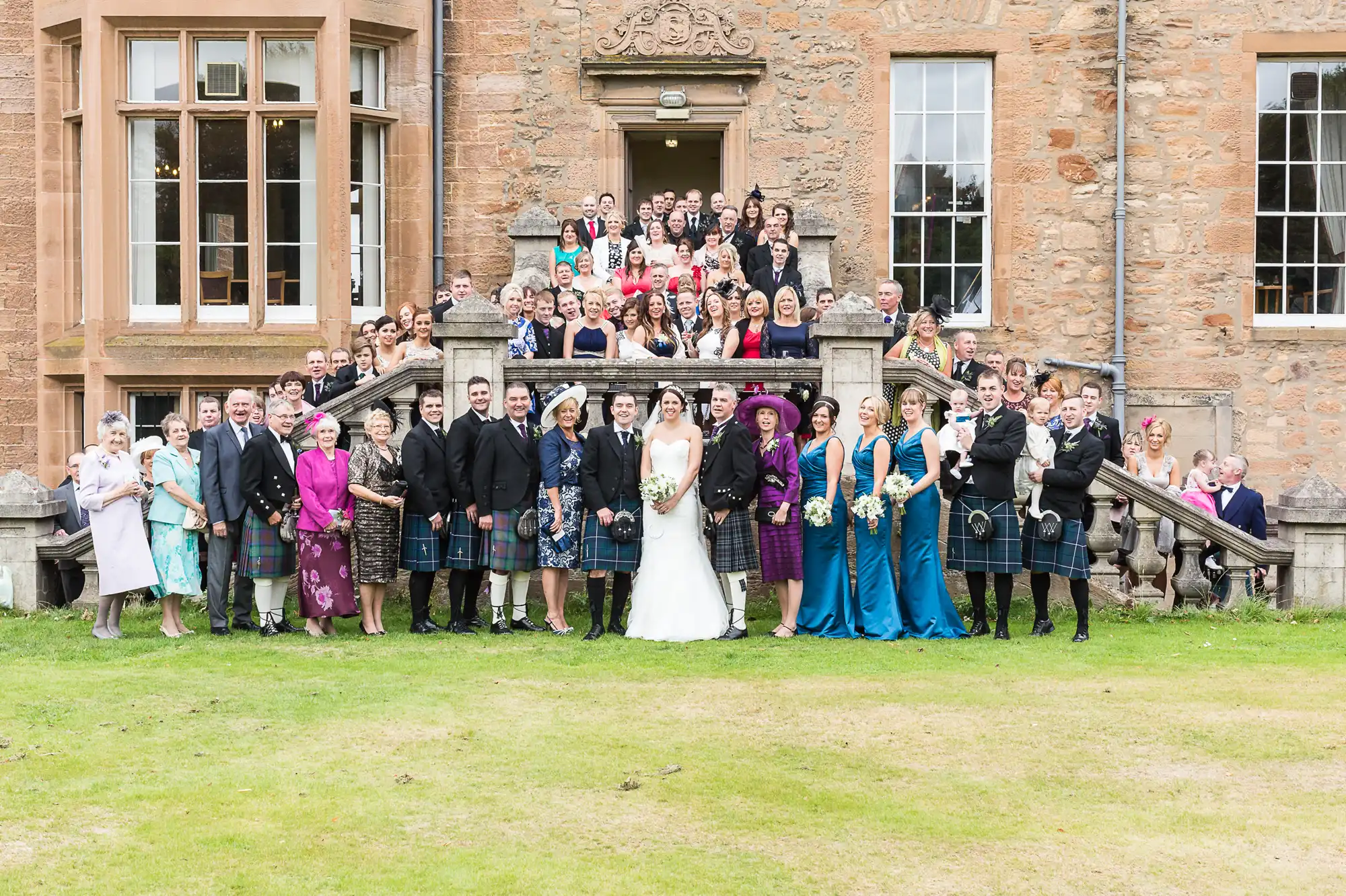 A large group of people in formal and traditional scottish attire posing for a photo at a wedding outside a historic brick building.