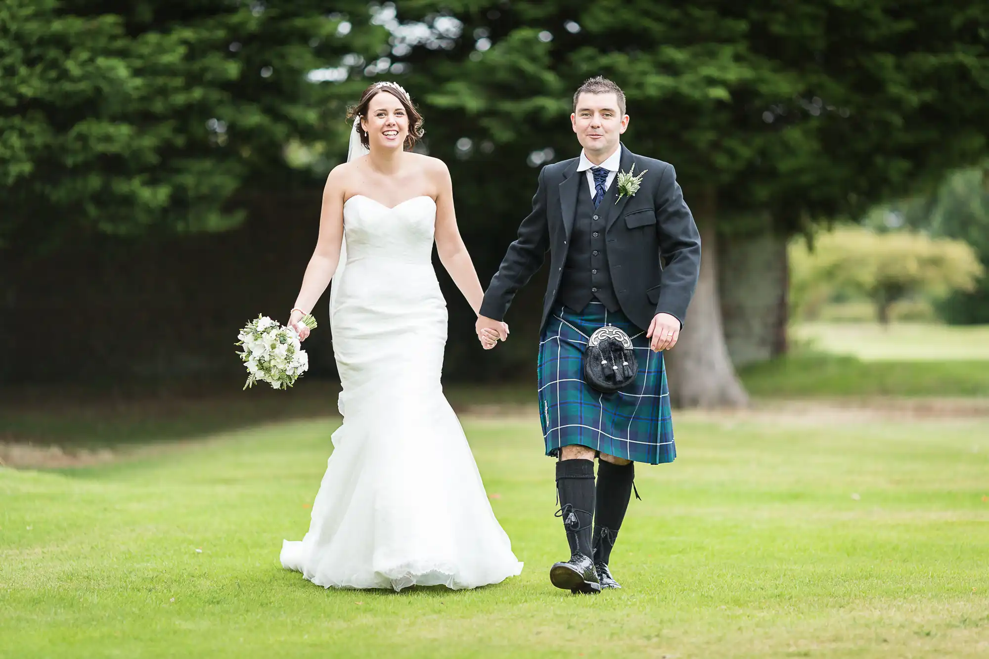 Bride in a white dress and groom in a kilt walking on grass, smiling, with trees in the background.