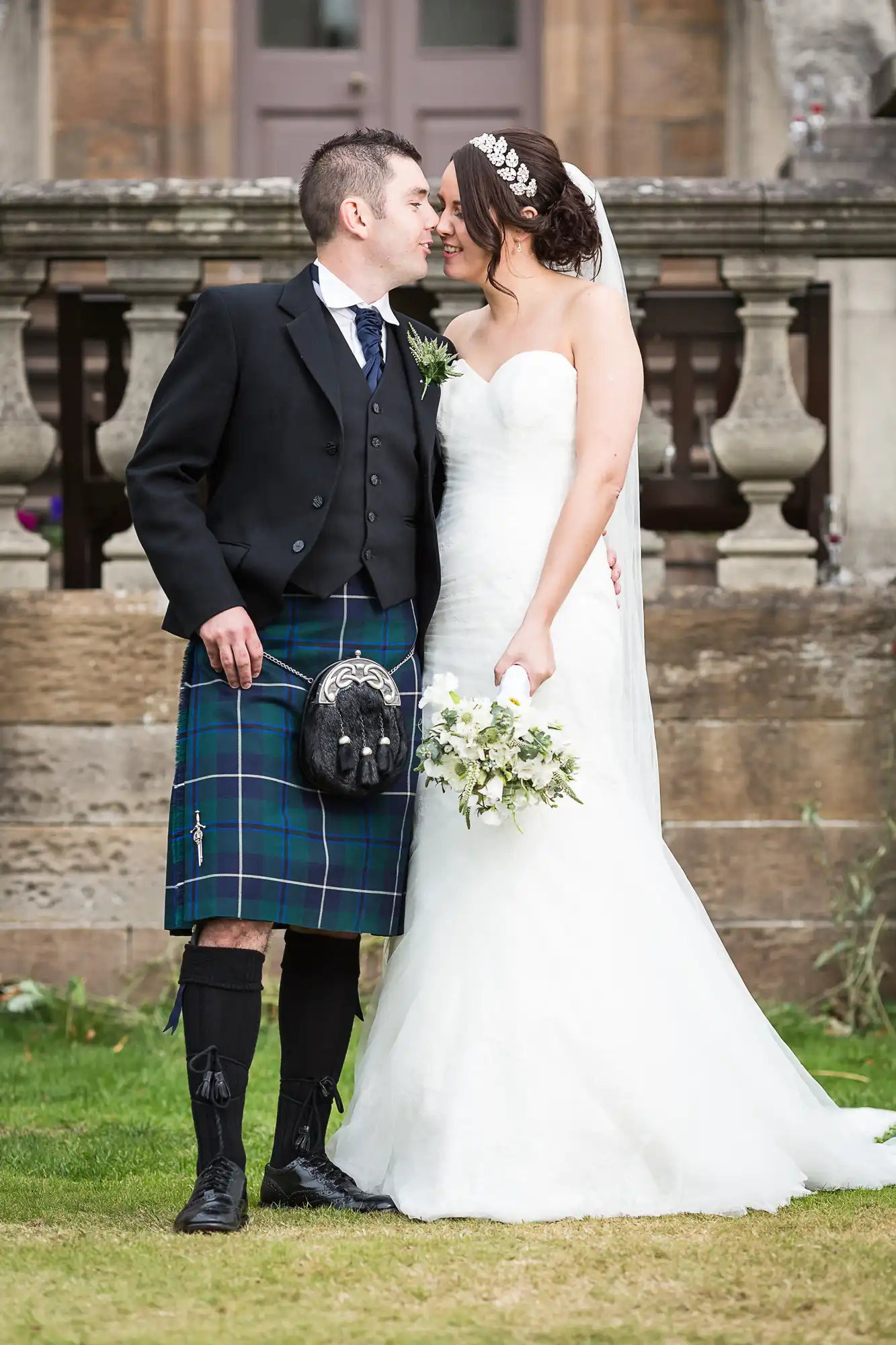 A bride in a white dress and a groom in a kilt embracing and smiling at each other in a garden.
