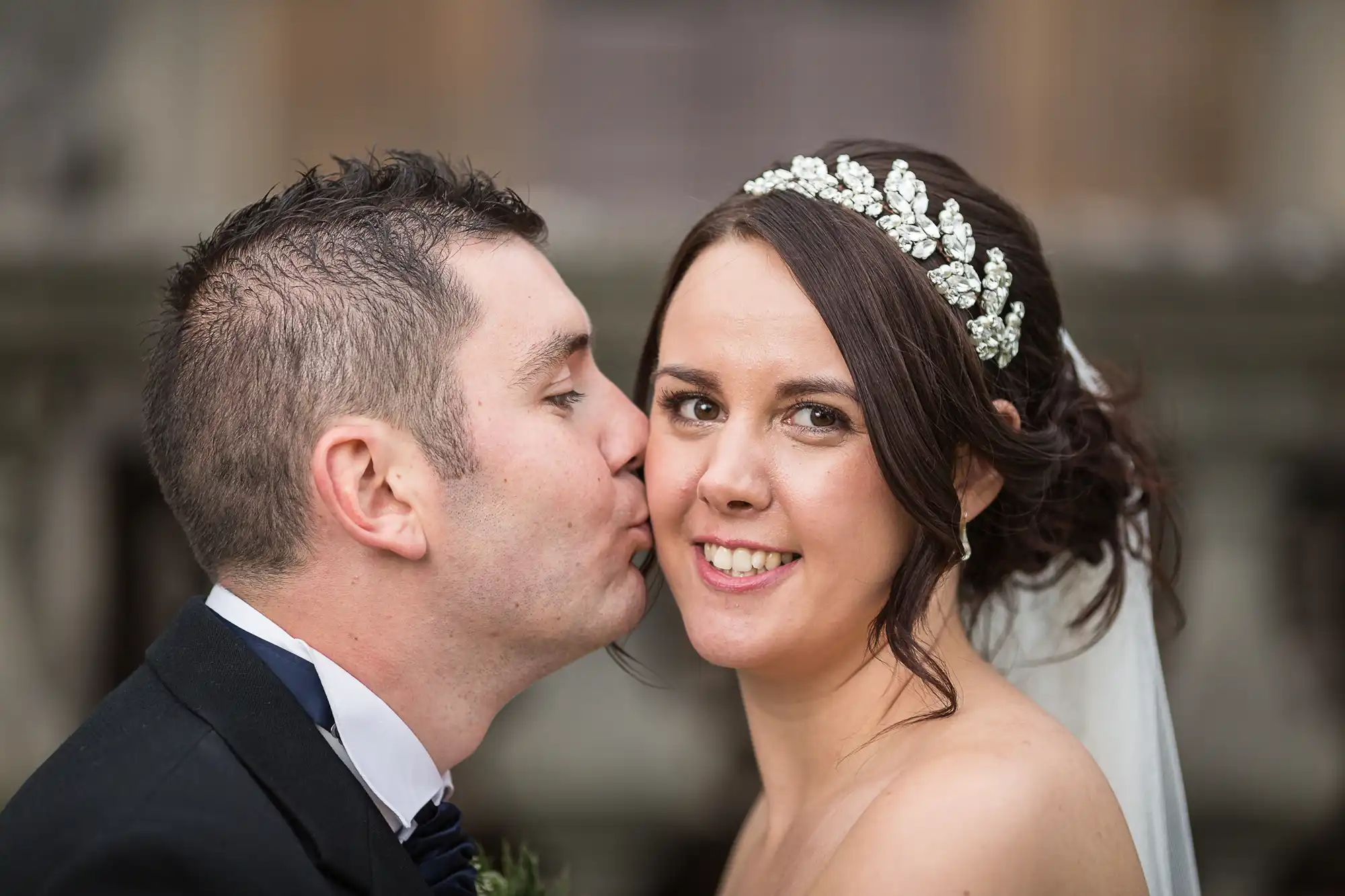 A groom in a black suit kisses the cheek of a bride wearing a white dress and a crystal tiara, with a soft-focus background.