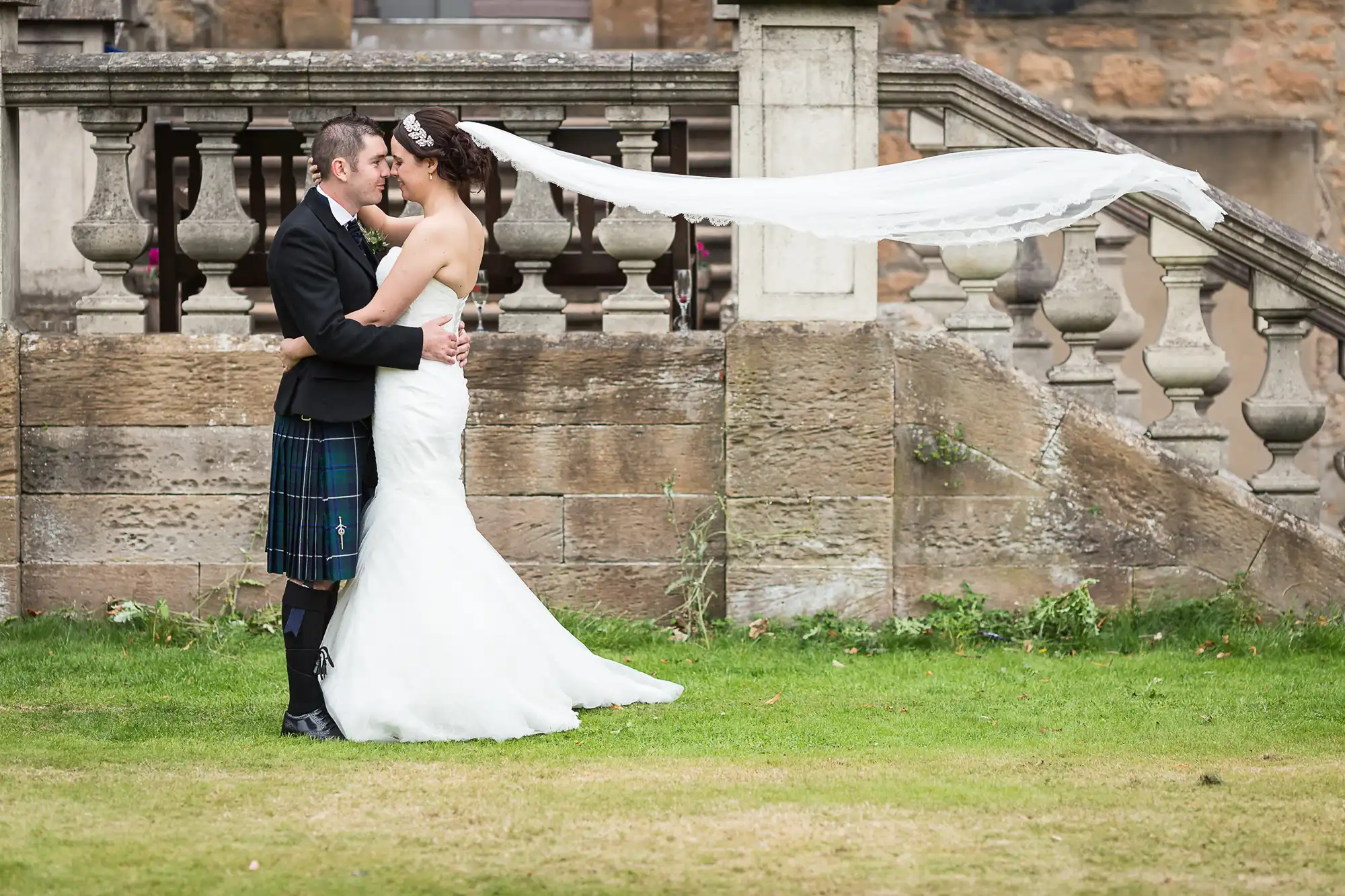 Bride in a white dress and groom in a kilt kissing, with the bride's veil blowing in the wind, standing next to a stone balustrade.