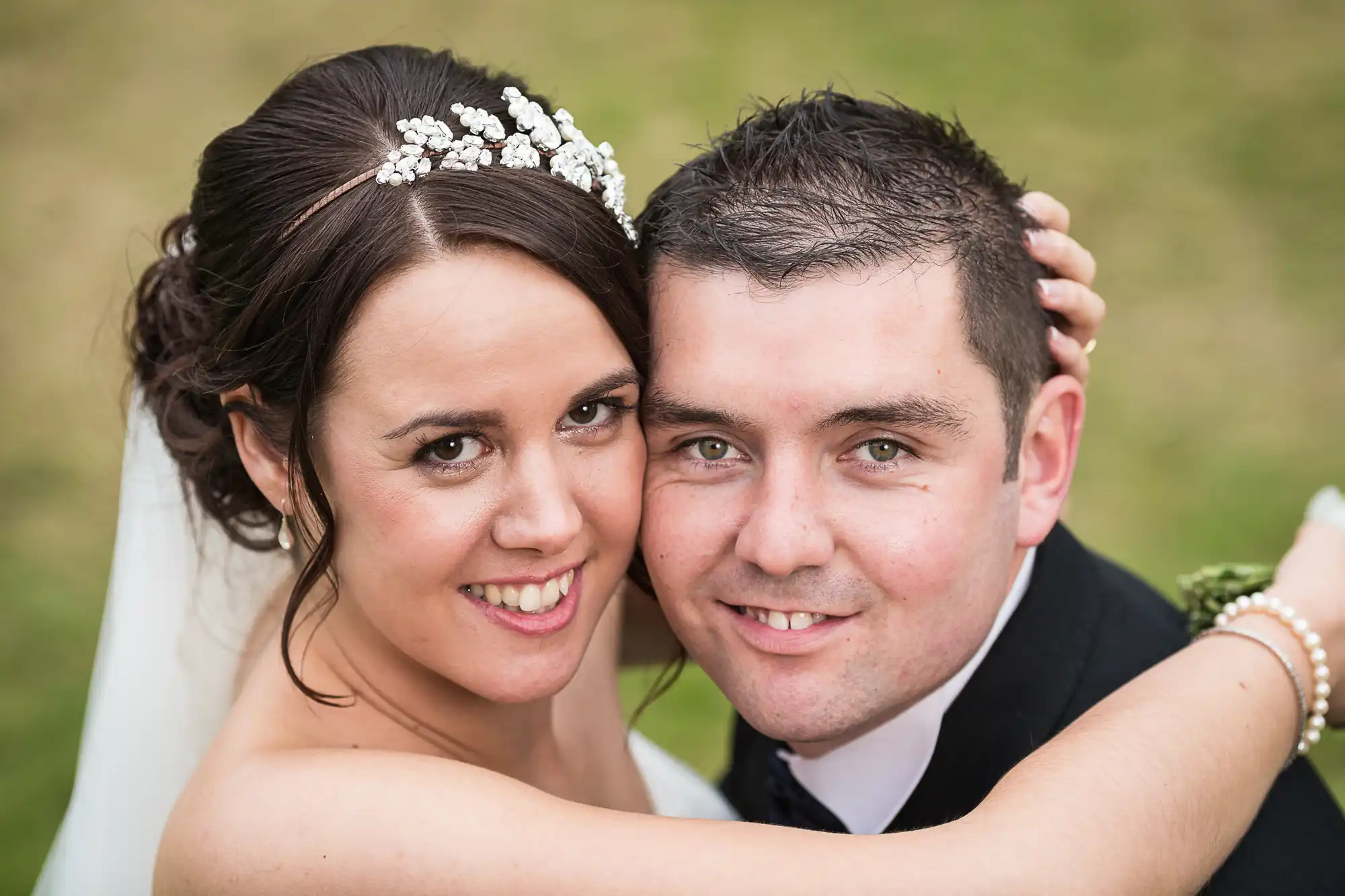 Close-up portrait of a smiling bride and groom outdoors, the bride wearing a tiara and veil, the groom in a black suit.