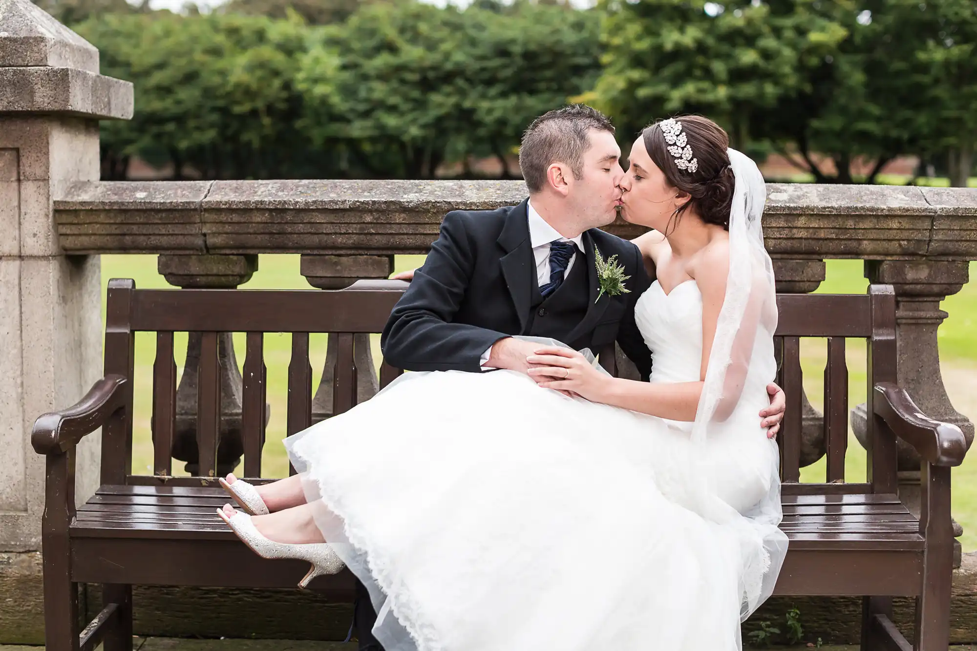 A bride in a white gown and a groom in a black suit kissing on a park bench, with trees in the background.