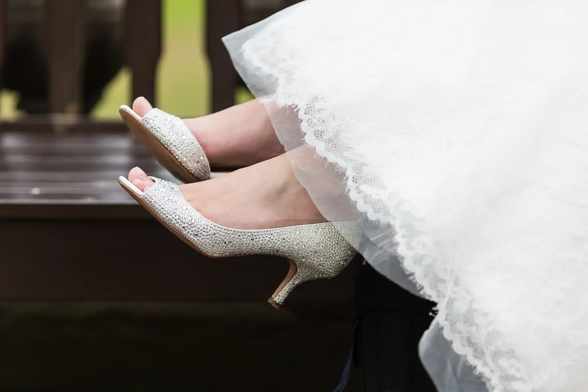 Bride wearing sparkly silver high heels, peeking from under a white lace wedding dress, seated on a bench.