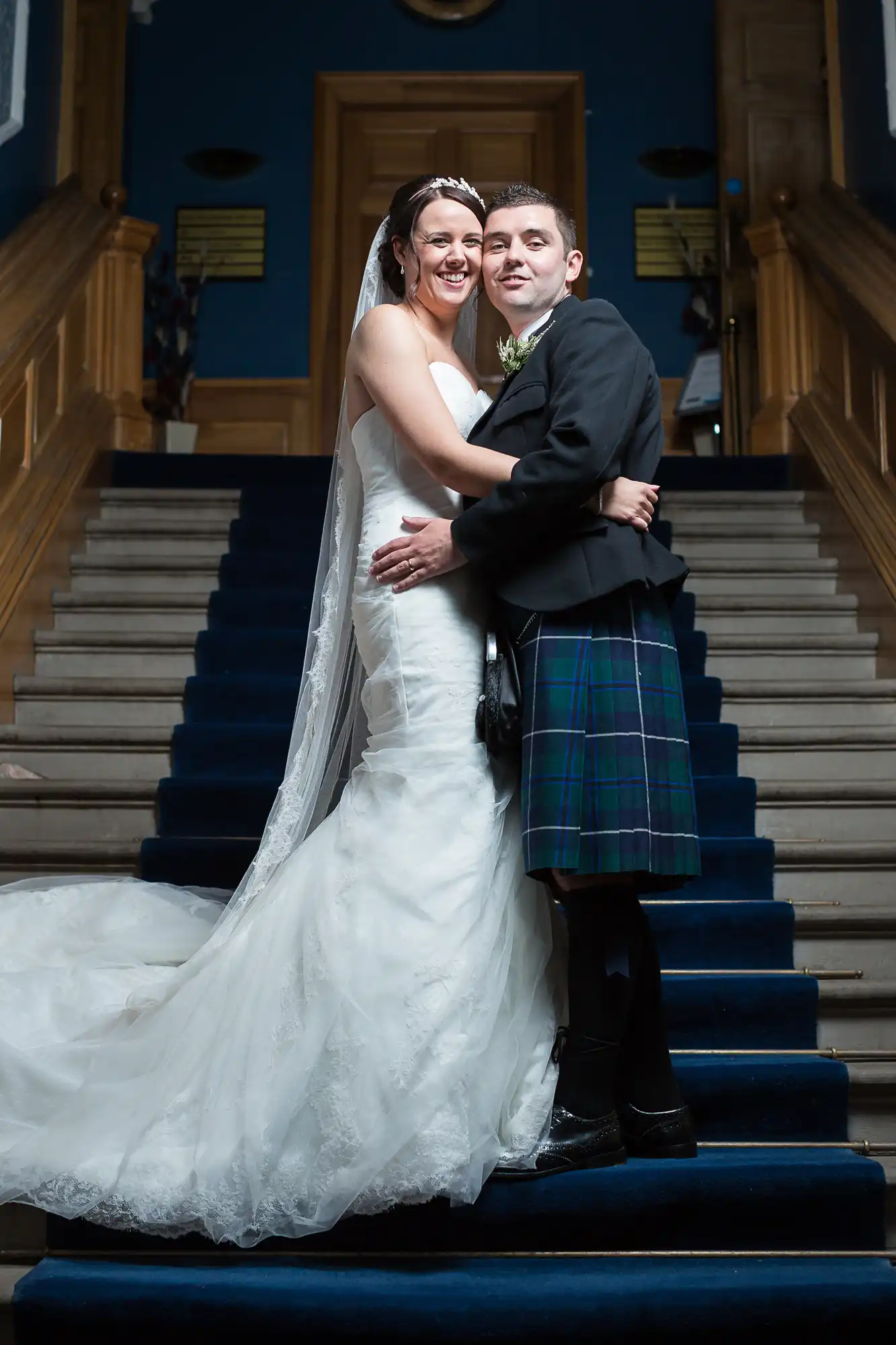 A bride in a white gown and a groom in a kilt embrace on a staircase, smiling joyfully at their wedding.