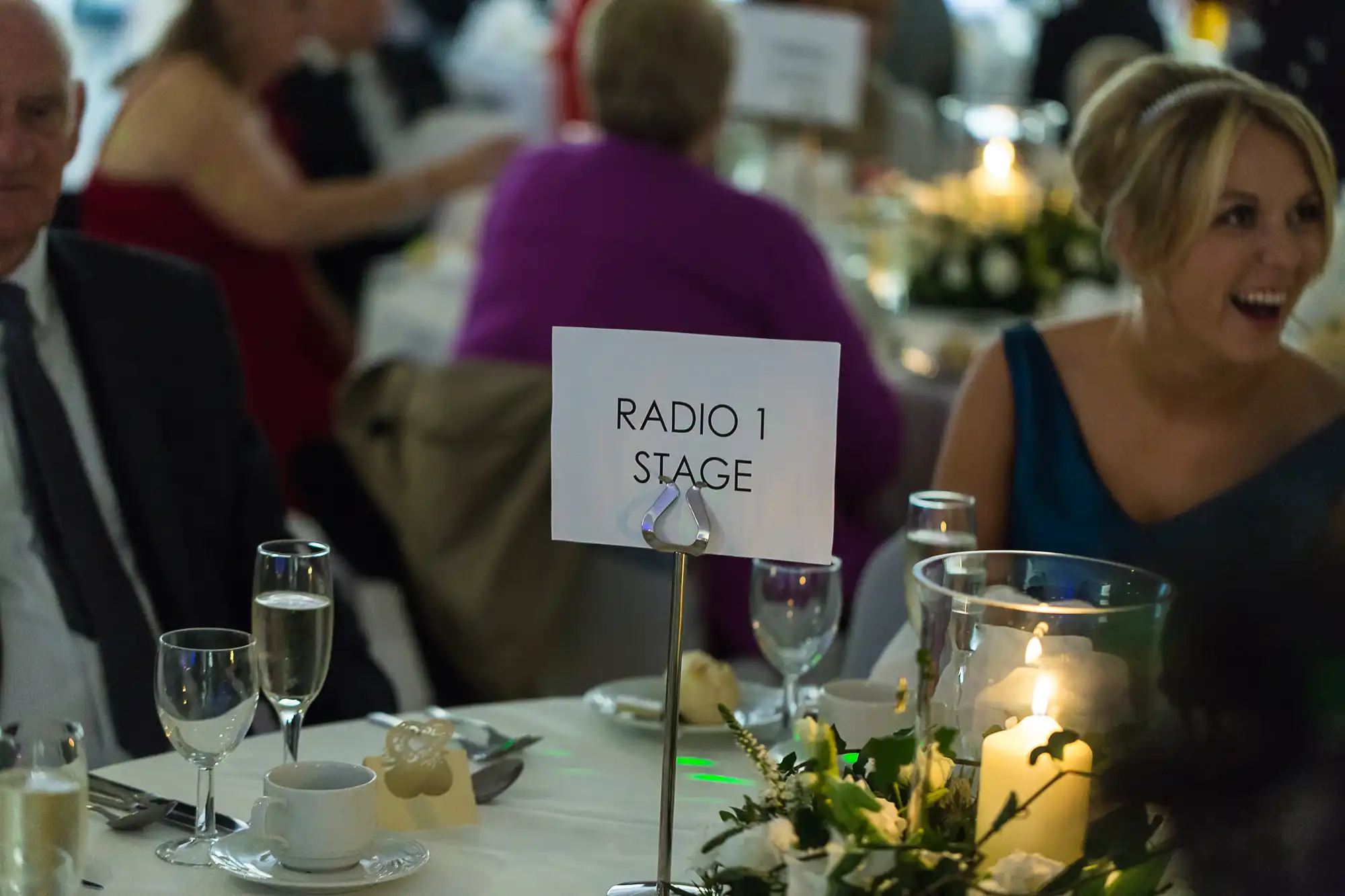 Table at a formal event with a sign reading "radio 1 stage," surrounded by guests and decorated with candles and a floral centerpiece.