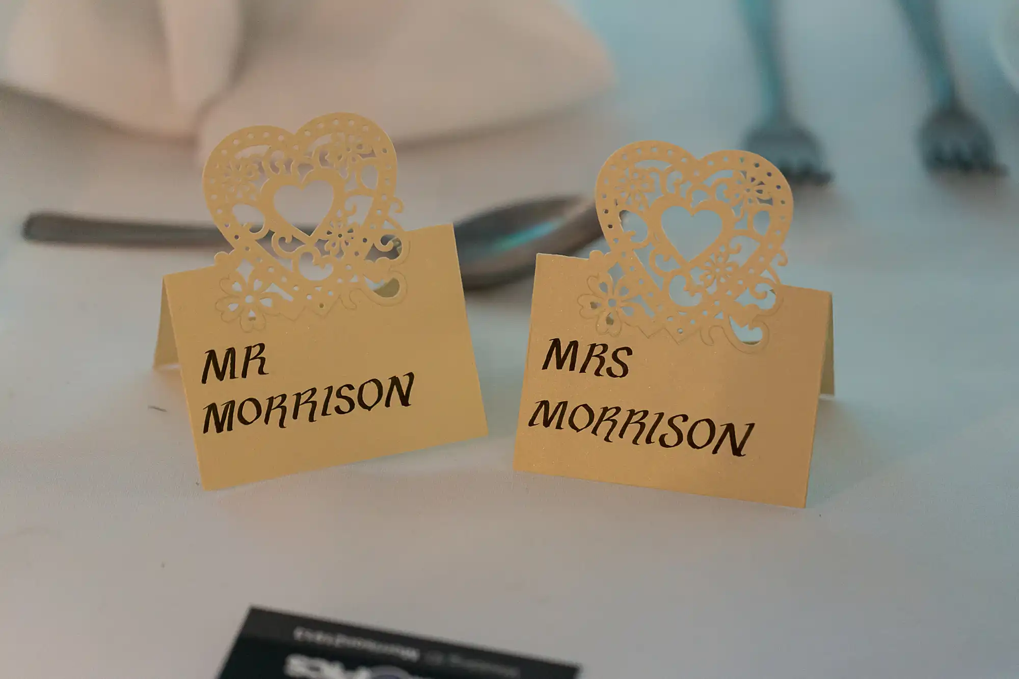 Two ornate wedding place cards reading "mr morrison" and "mrs morrison" on a table, with soft focus on utensils in the background.