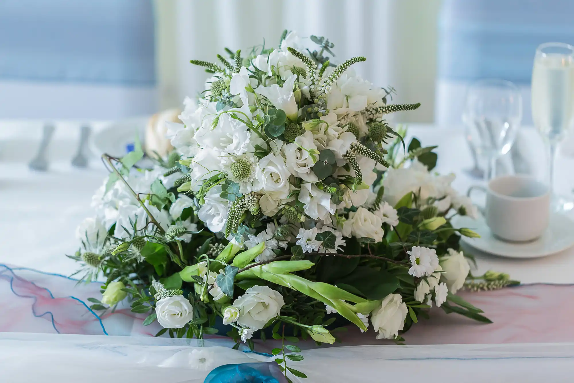 A lush bouquet of white flowers with green foliage on a table, next to a glass of champagne and a coffee cup.