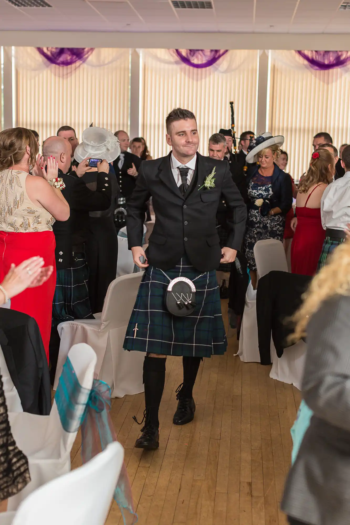 A man in a kilt and jacket smiling as he walks through a clapping crowd at a lively indoor event.