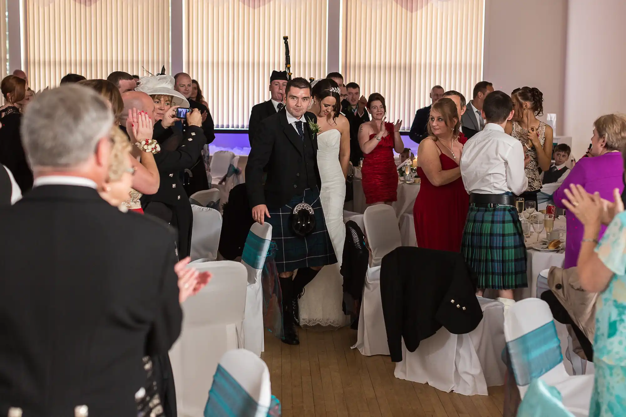 A bride and groom walking through a clapping crowd at a scottish wedding reception, with the groom wearing a kilt.