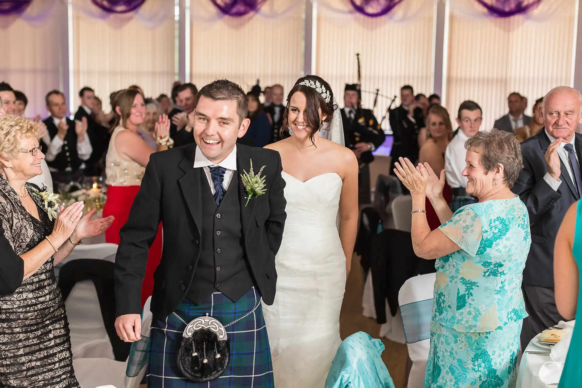 A bride and groom walk through a clapping crowd in a decorated hall, the groom wearing a kilt and the bride in a white dress.