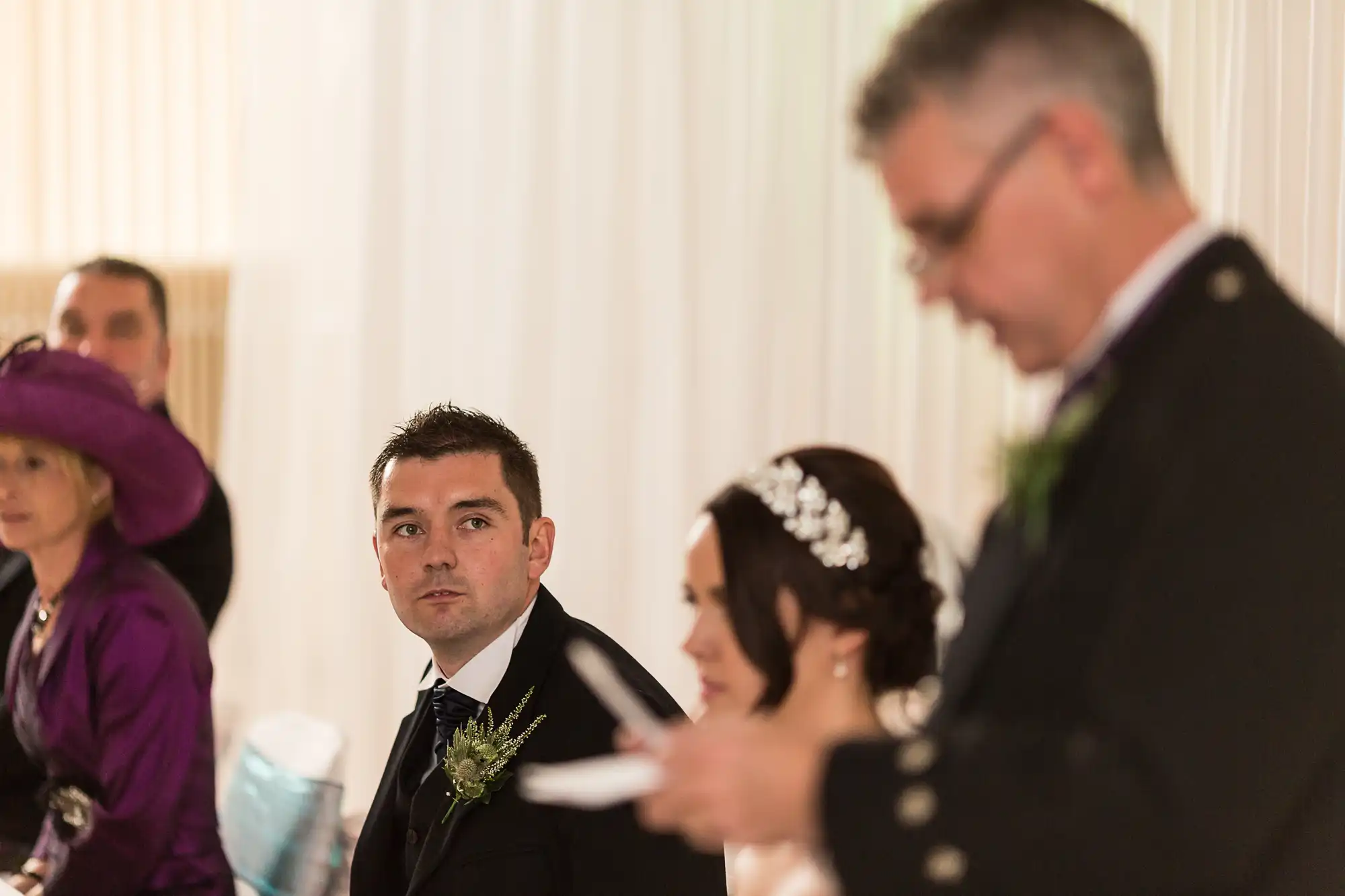 A groom in a black suit looks intently ahead during a wedding ceremony, with an officiant and wedding guests slightly blurred in the foreground and background.