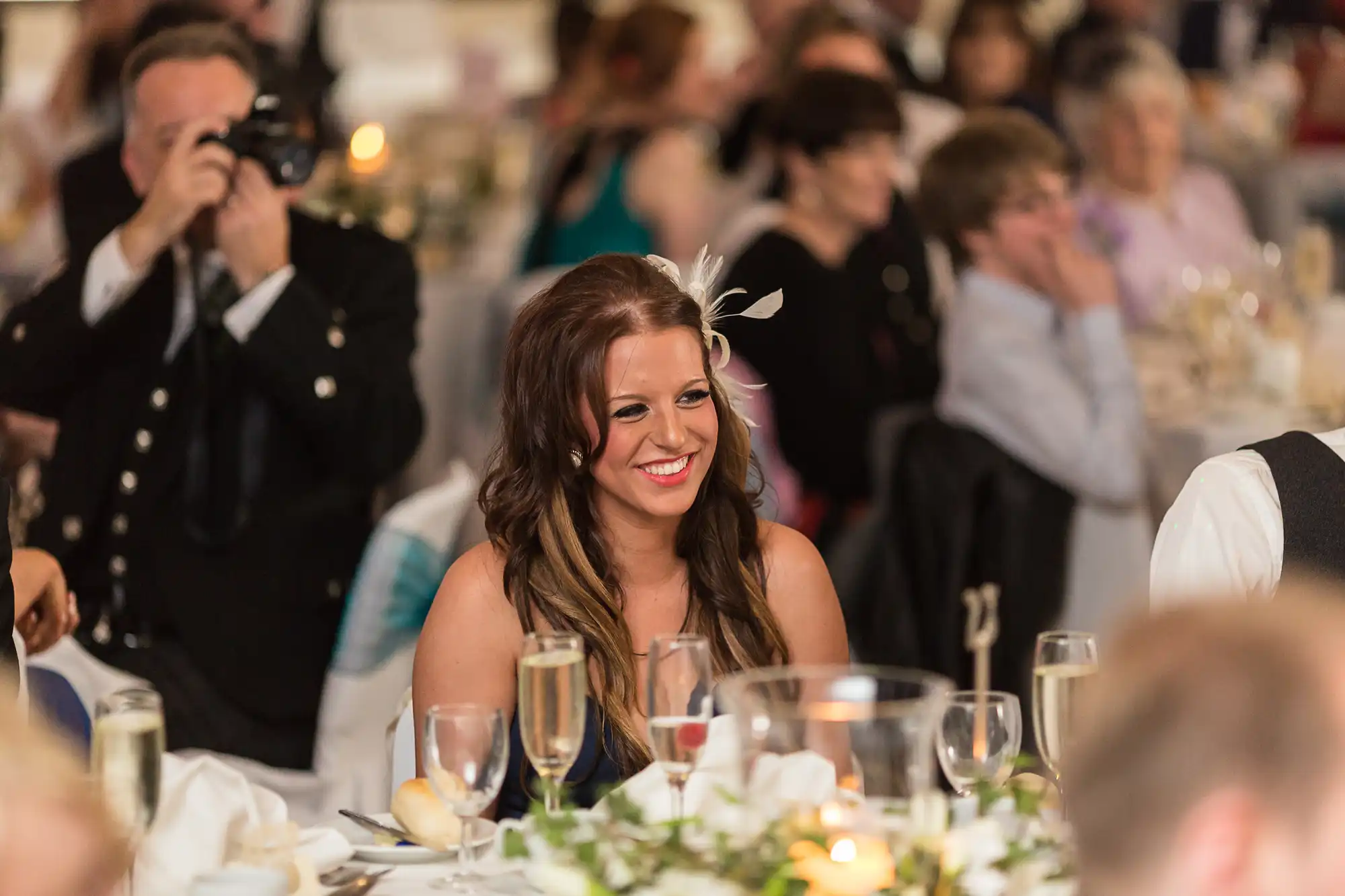 A woman with a feathered hairpiece smiling at a banquet table during an event, with guests and a photographer in the background.