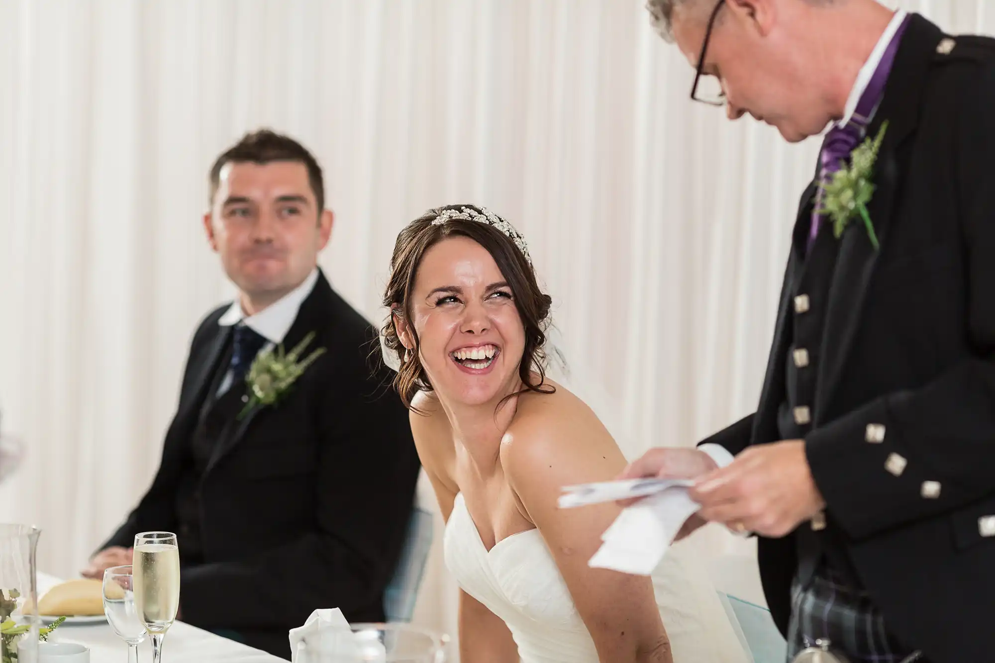 A bride laughing joyously at a wedding reception table as a man presents something, with a groom smiling in the background.