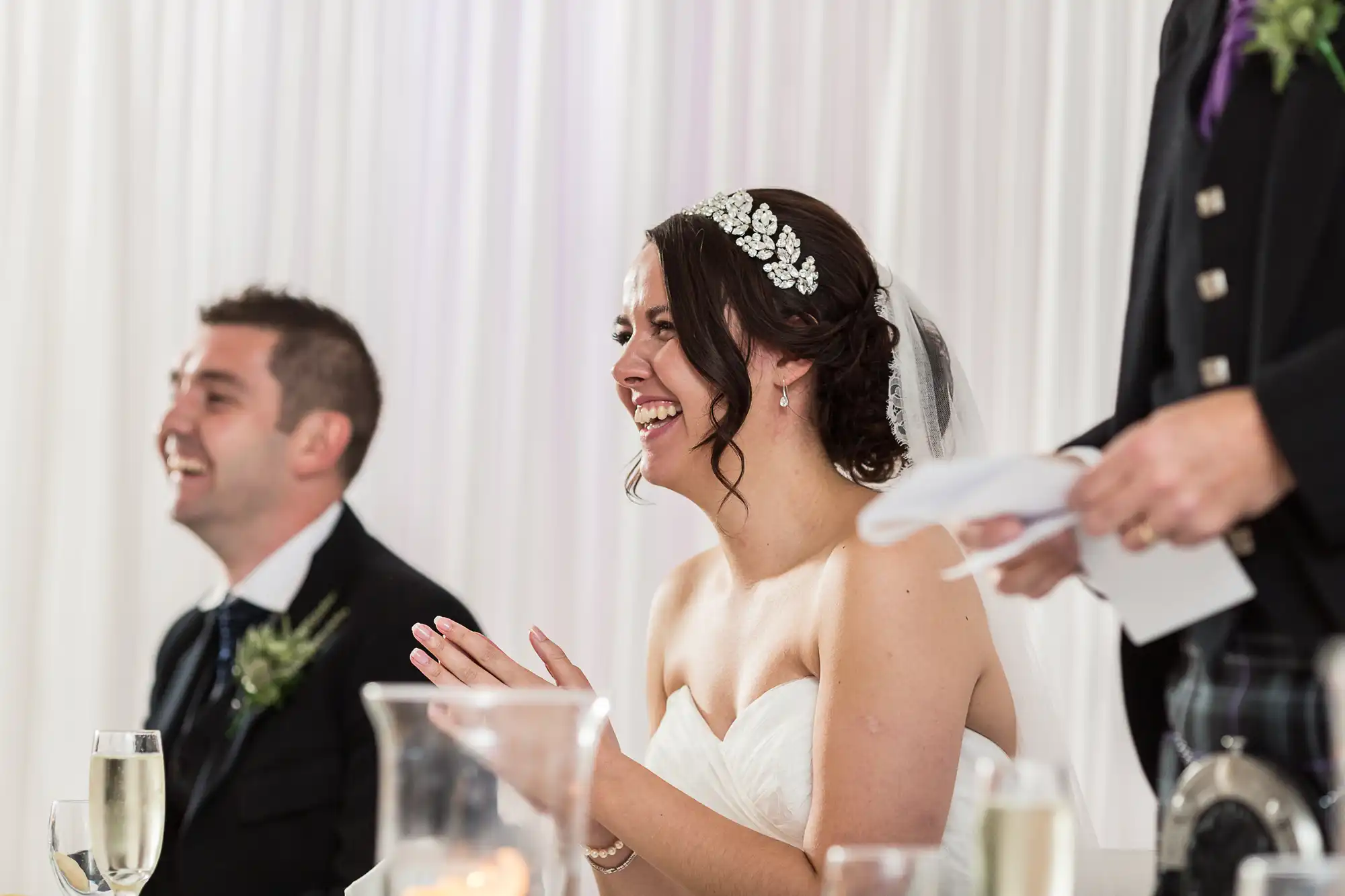 Bride laughing joyfully at a wedding reception table, wearing a white dress and floral headpiece, with a smiling groom in the background.