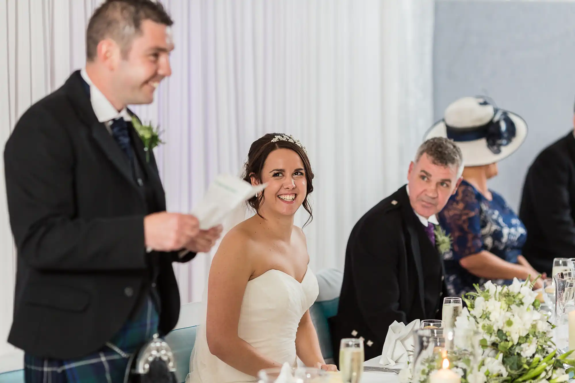 A bride in a white dress smiles at a wedding reception, while a man in a kilt holds a microphone next to her; other guests visible in the background.