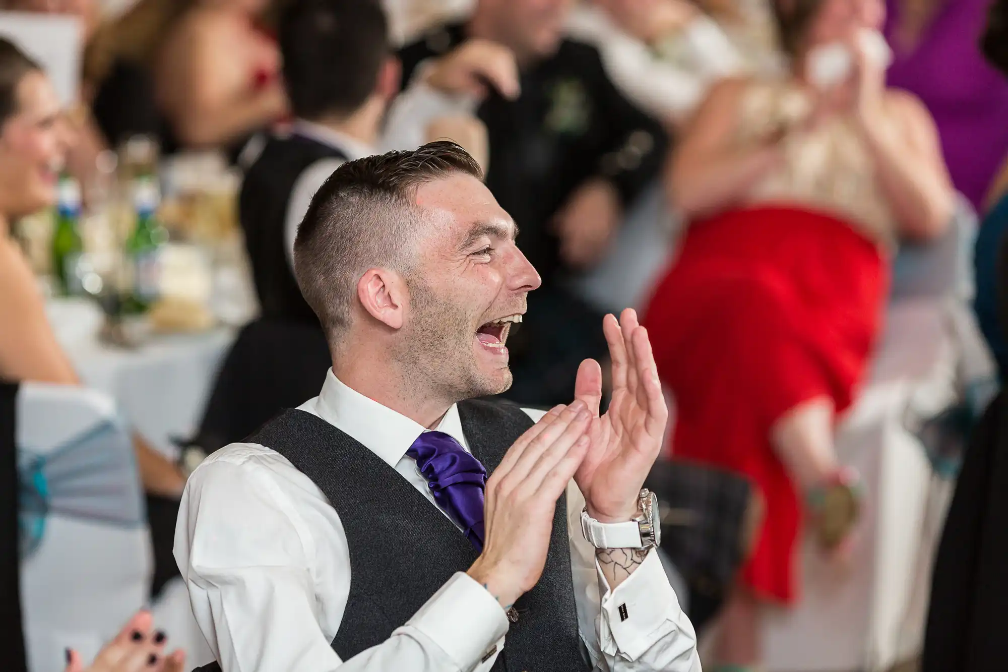 Man in a gray suit and purple tie clapping and laughing at a festive event with blurred guests in the background.
