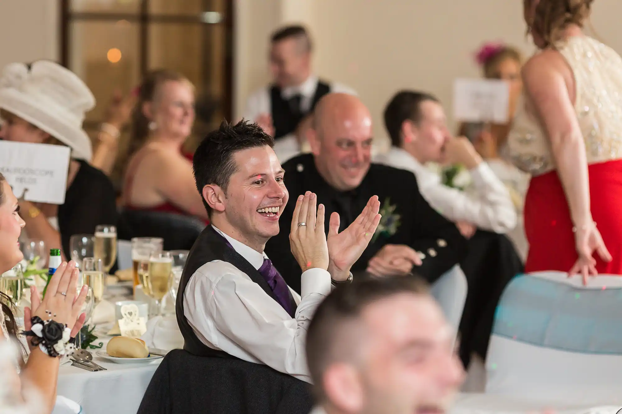 A man in a formal suit clapping and smiling at a wedding reception, surrounded by other guests at decorated tables.