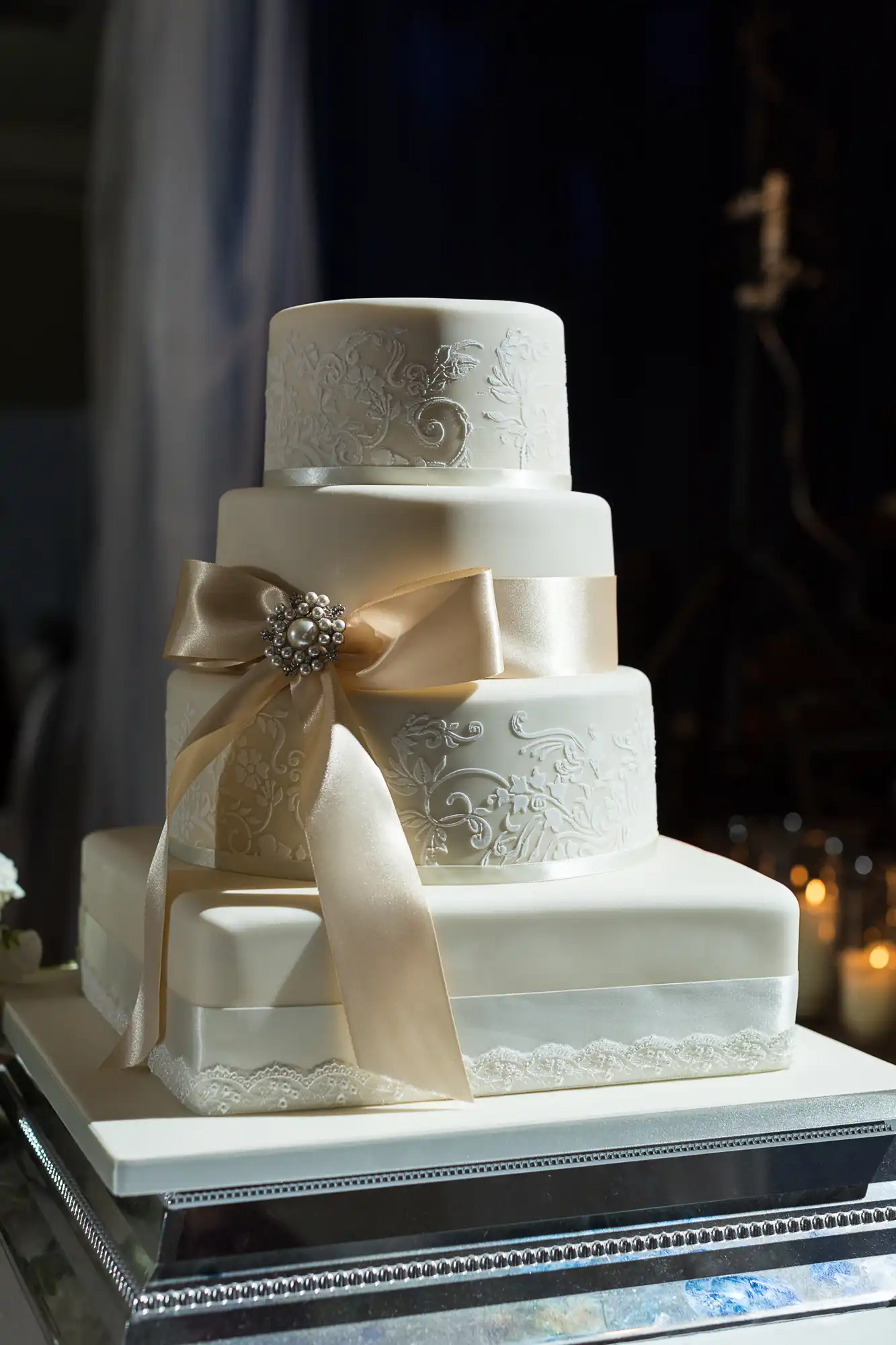 A four-tiered white wedding cake decorated with ornate patterns and a satin ribbon, topped with a jeweled brooch, displayed on a silver stand.