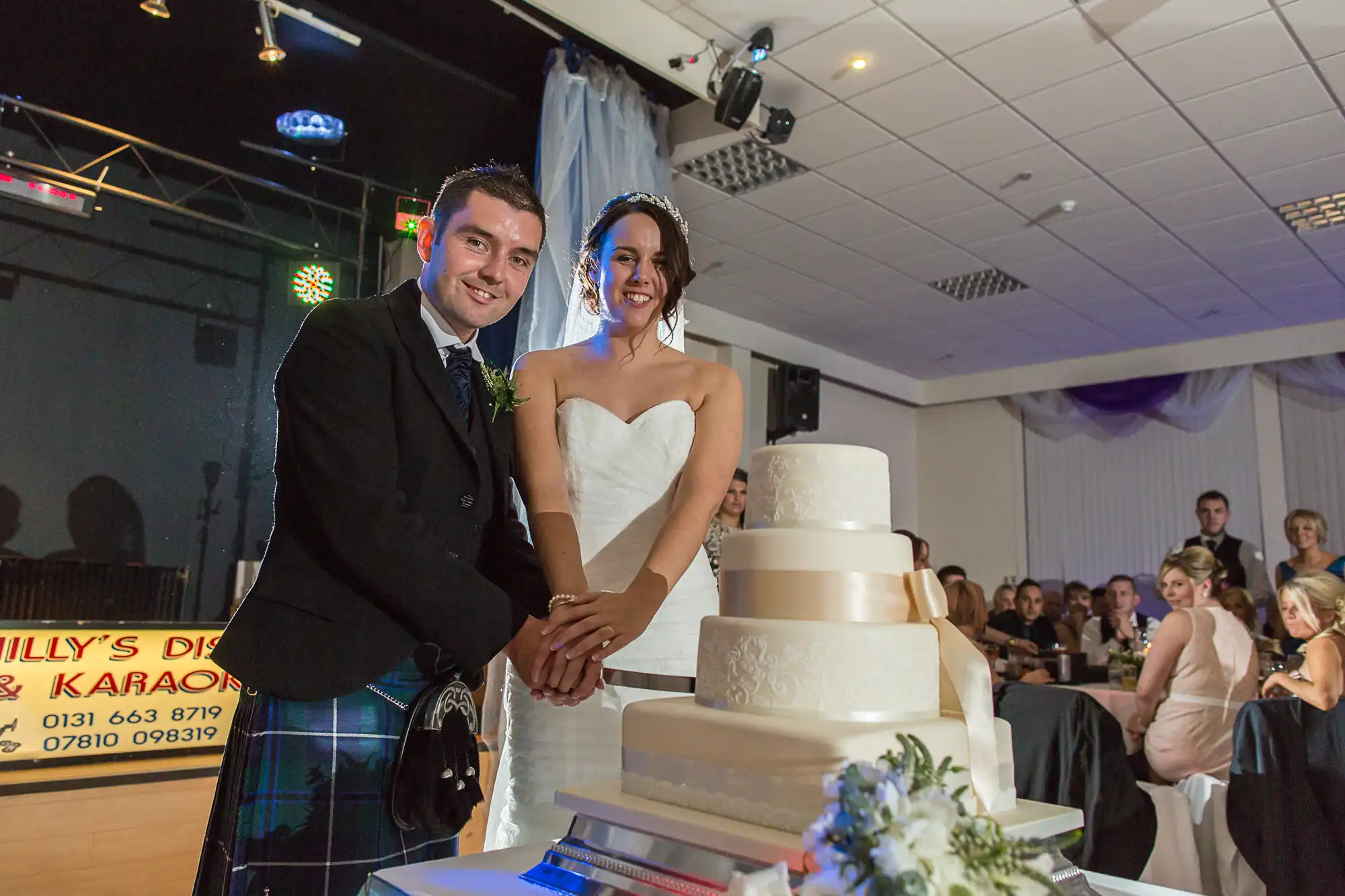 A bride and groom smiling at their wedding reception, standing behind a large tiered cake, with guests and a dj booth in the background.