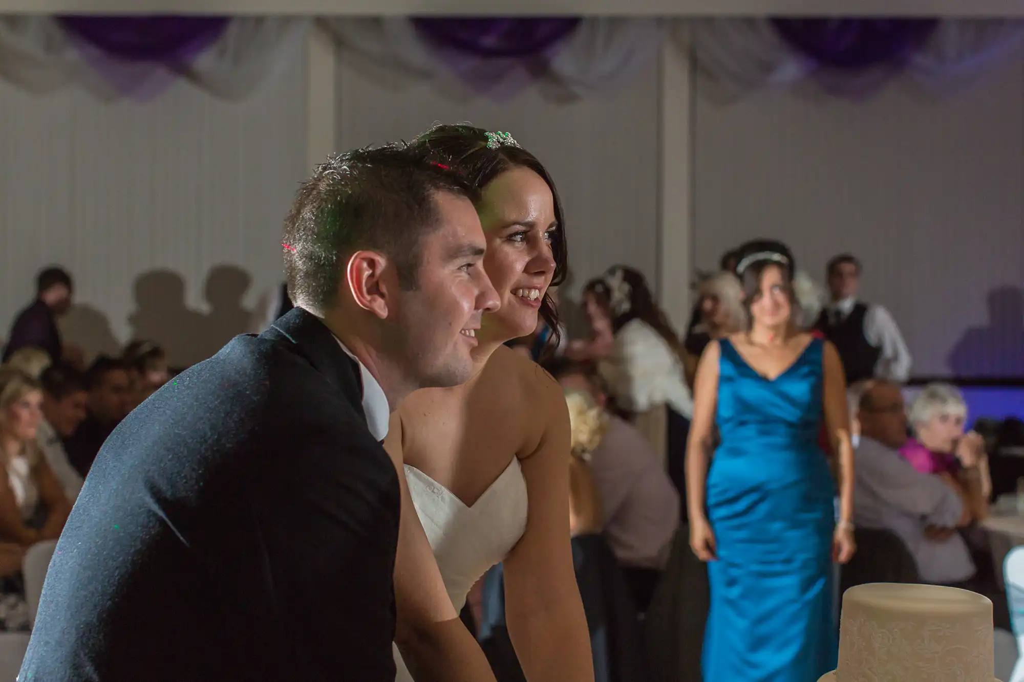 A bride and groom, smiling and looking ahead, sitting during a wedding reception, with guests in the background.