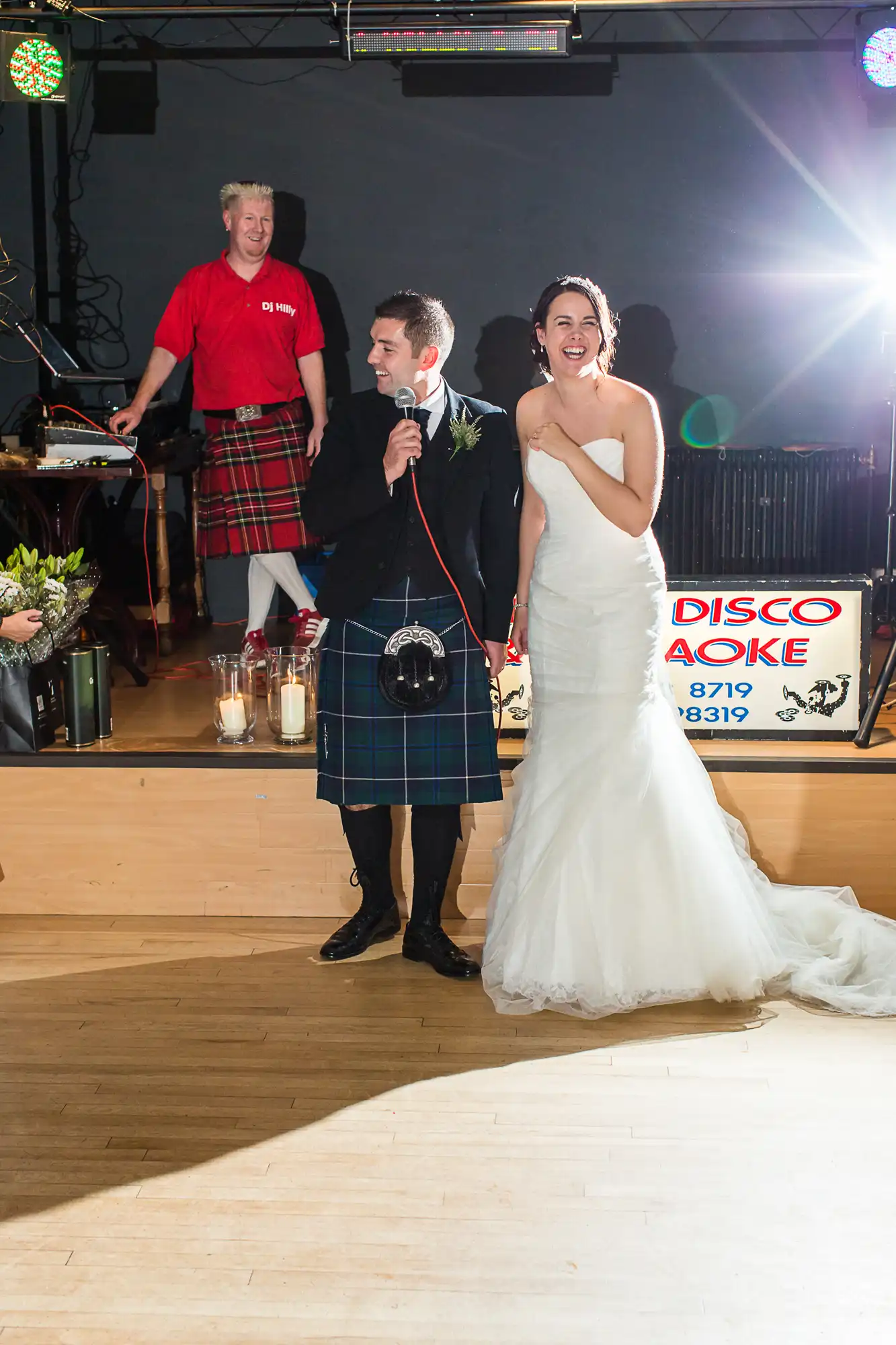 A bride and groom laugh joyously on a dance floor, with the groom holding a microphone. a man in a kilt observes behind them, next to a "disco karaoke" sign.