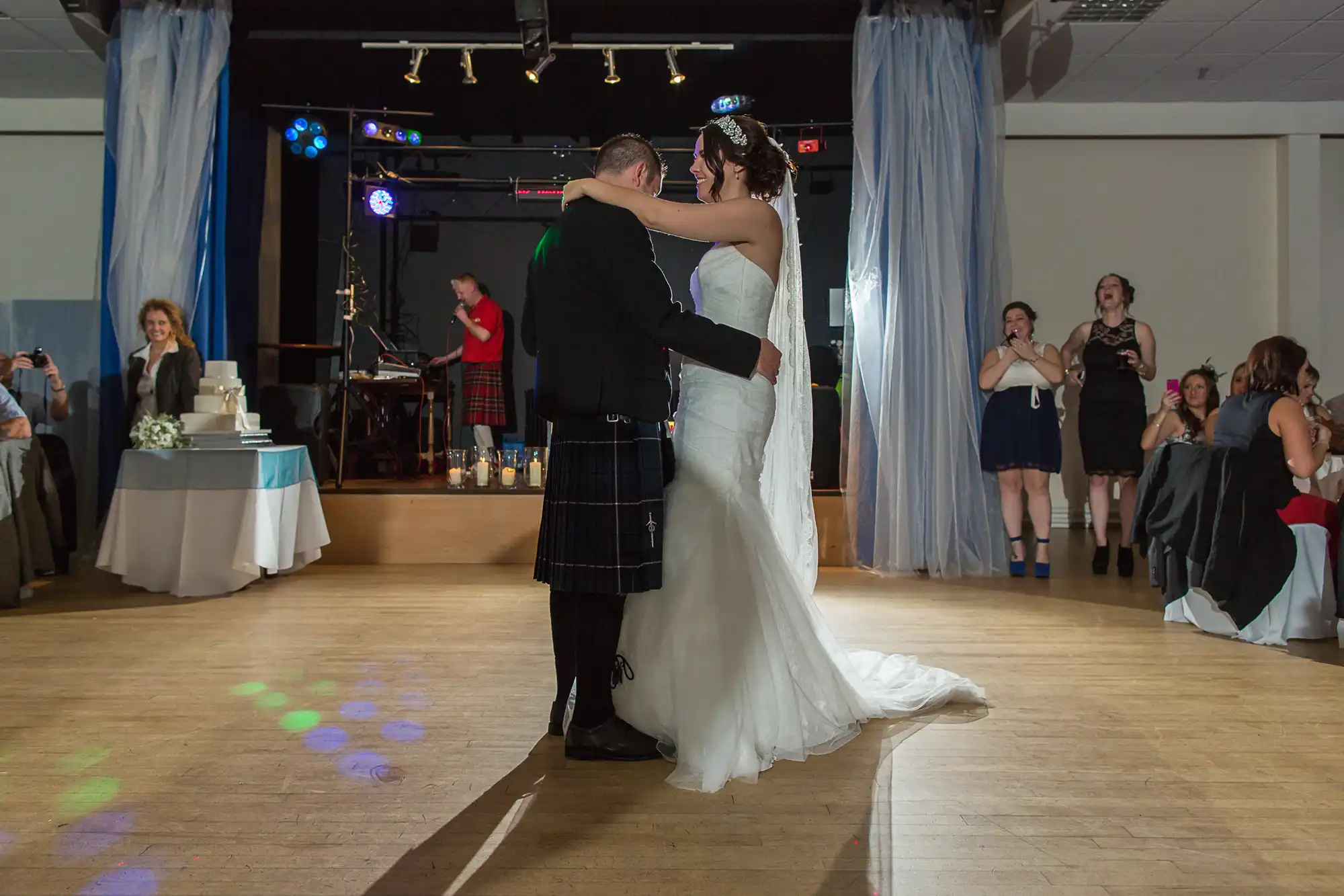 A bride and groom share their first dance at a wedding reception, with guests watching in a decorated hall. the groom wears a kilt; colorful lights dot the dance floor.