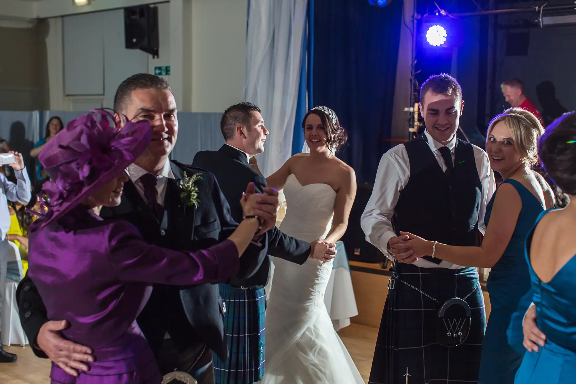 A joyful wedding reception with guests dancing; the bride and groom in the center, surrounded by guests in formal attire, including kilts.