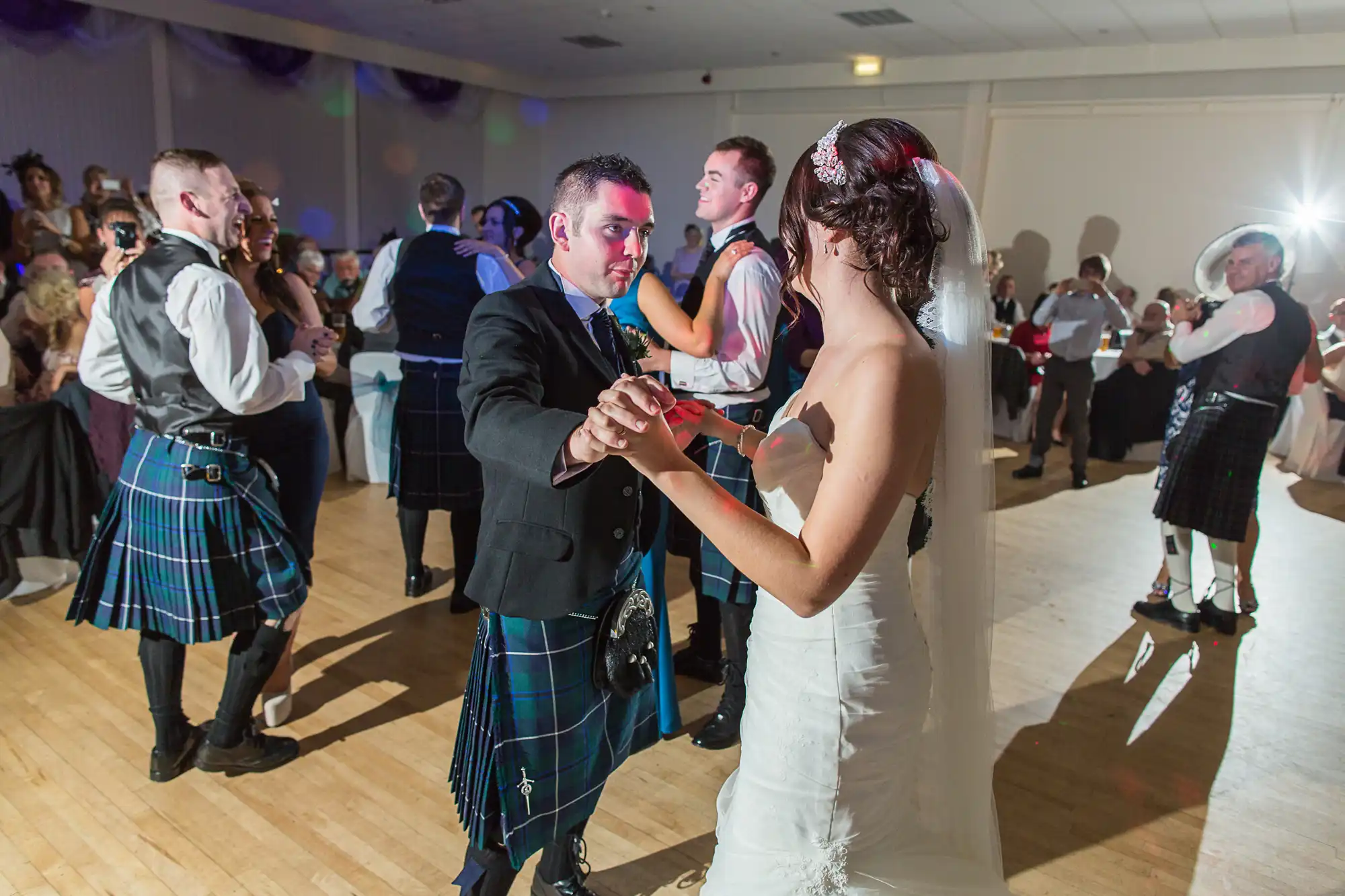 Bride and groom dancing at their wedding reception, surrounded by guests in scottish kilts and modern attire.