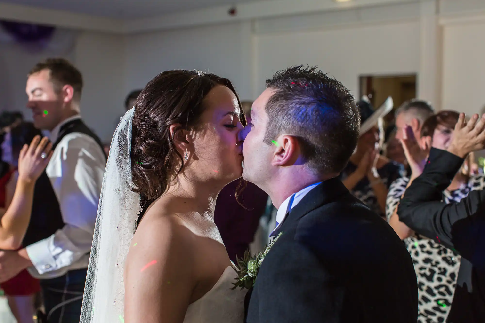 A bride and groom kiss on the dance floor surrounded by clapping guests during a wedding reception.