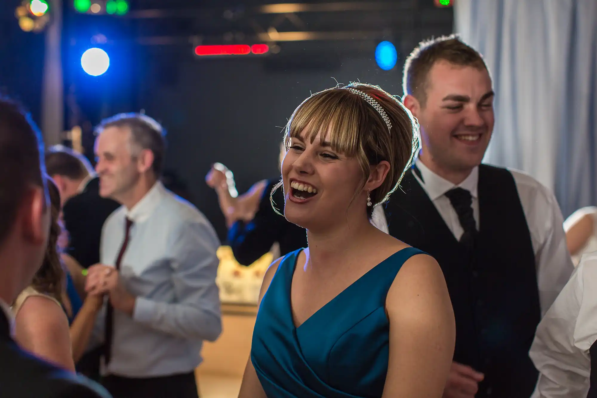 A joyful woman in a blue dress laughs while dancing at a lively party with people in the background.