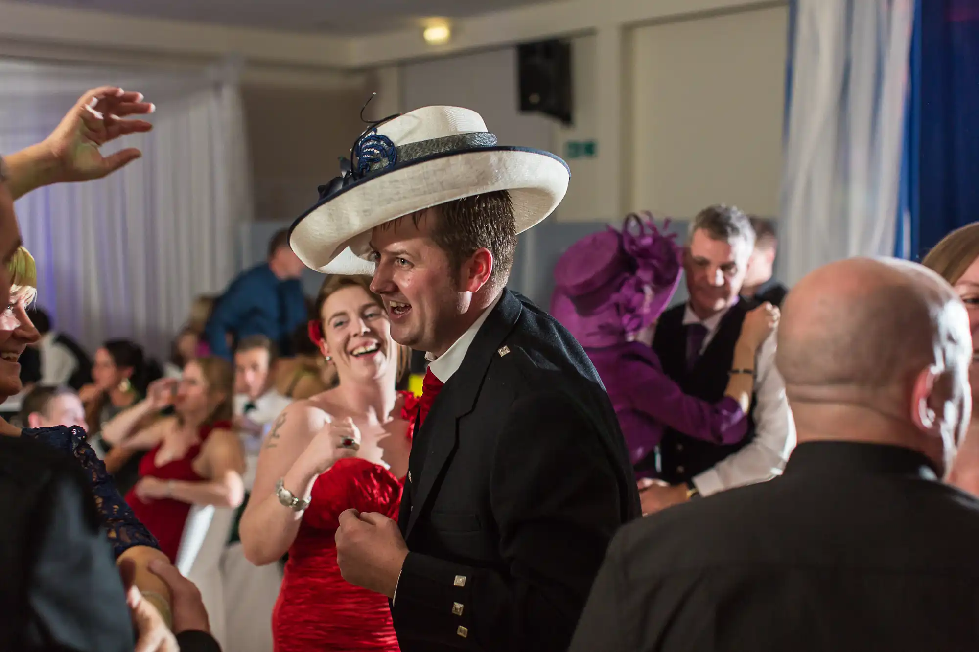 A man in a suit and cowboy hat dances with a woman in a red dress at a lively indoor wedding reception, surrounded by smiling guests.