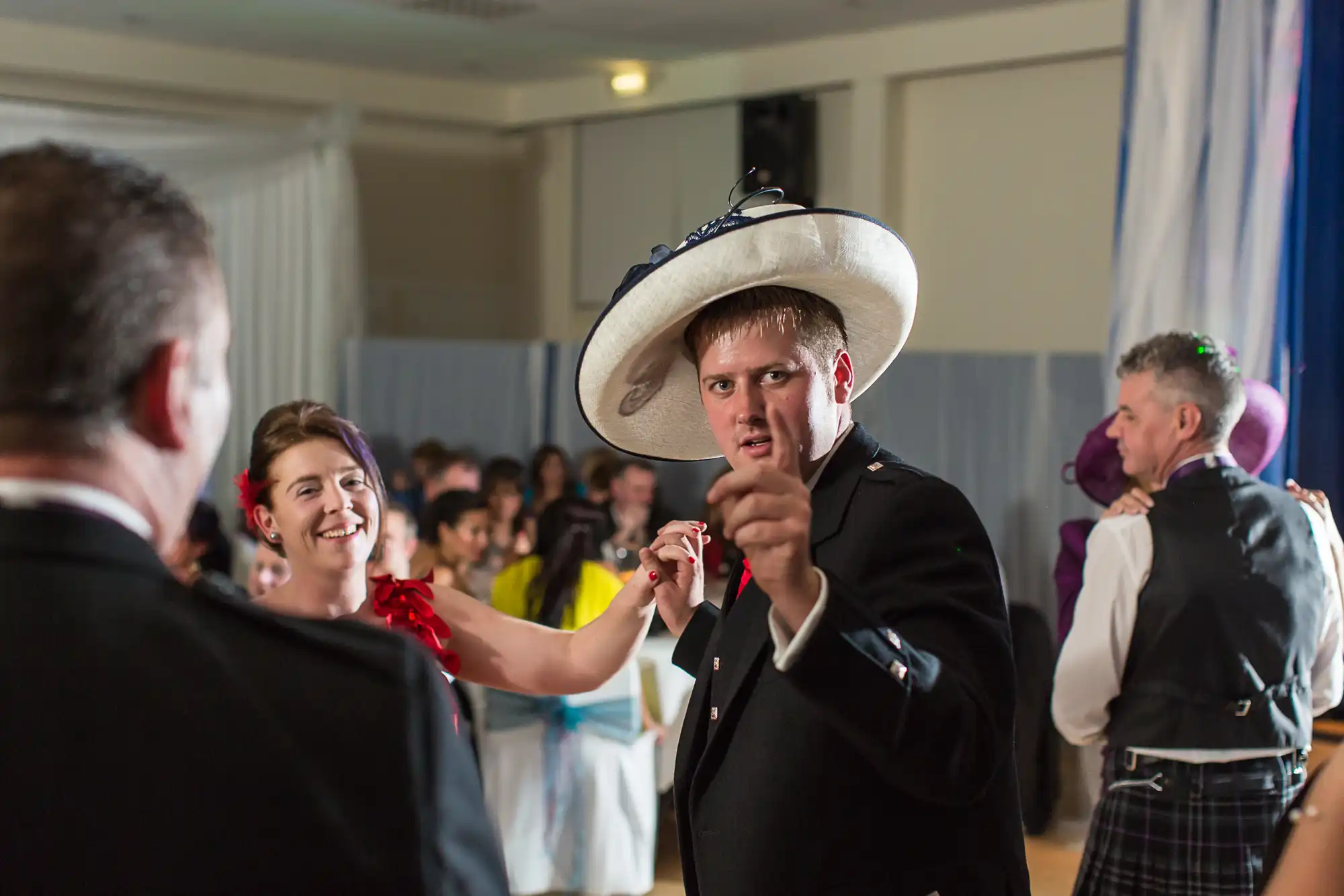 A man in a suit and oversized hat points and looks directly at the camera during a lively dance at a festive event, surrounded by other guests.