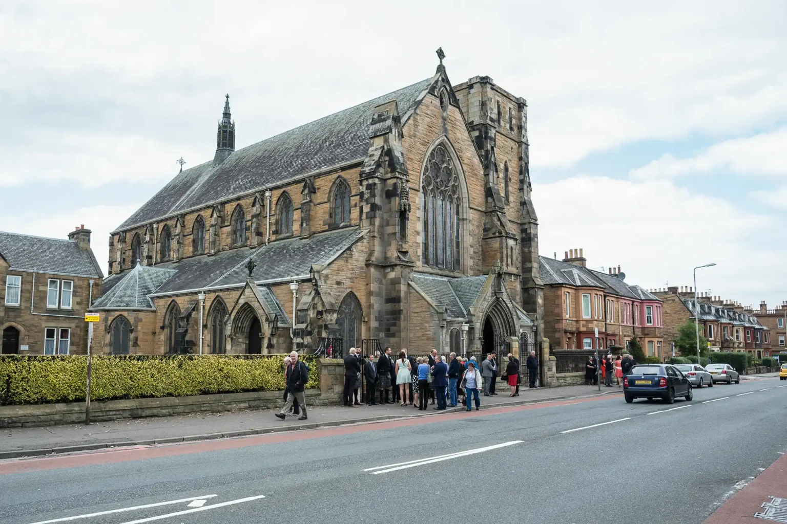A group of people standing outside a large gothic-style church on a street, with cars passing by and residential buildings in the background.