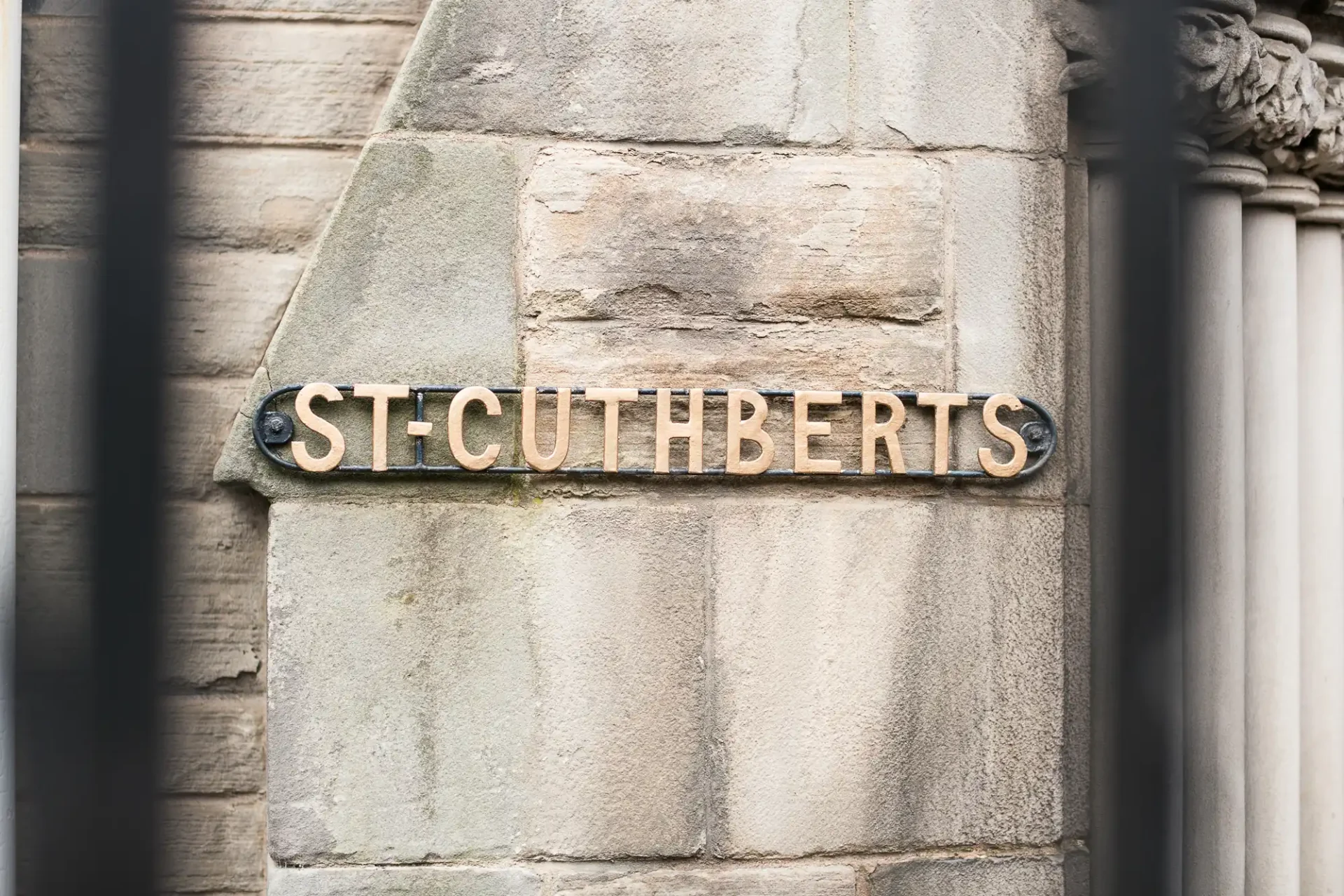 A metal plaque reading "stcuthberts" mounted on a stone wall, viewed through blurred vertical bars in the foreground.