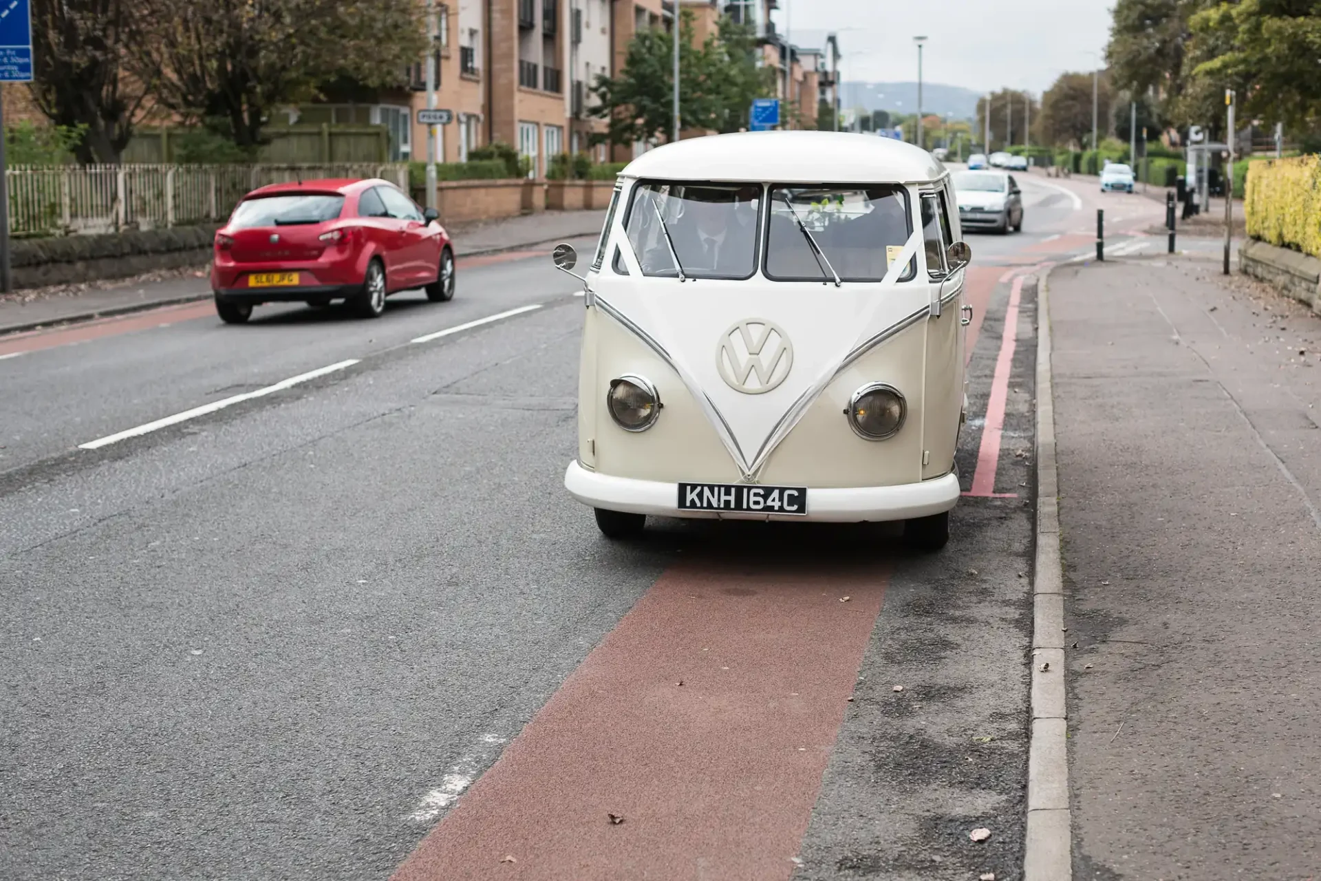 A vintage white volkswagen van parked on a city street with a red bike lane beside it. other cars and buildings are visible in the background.
