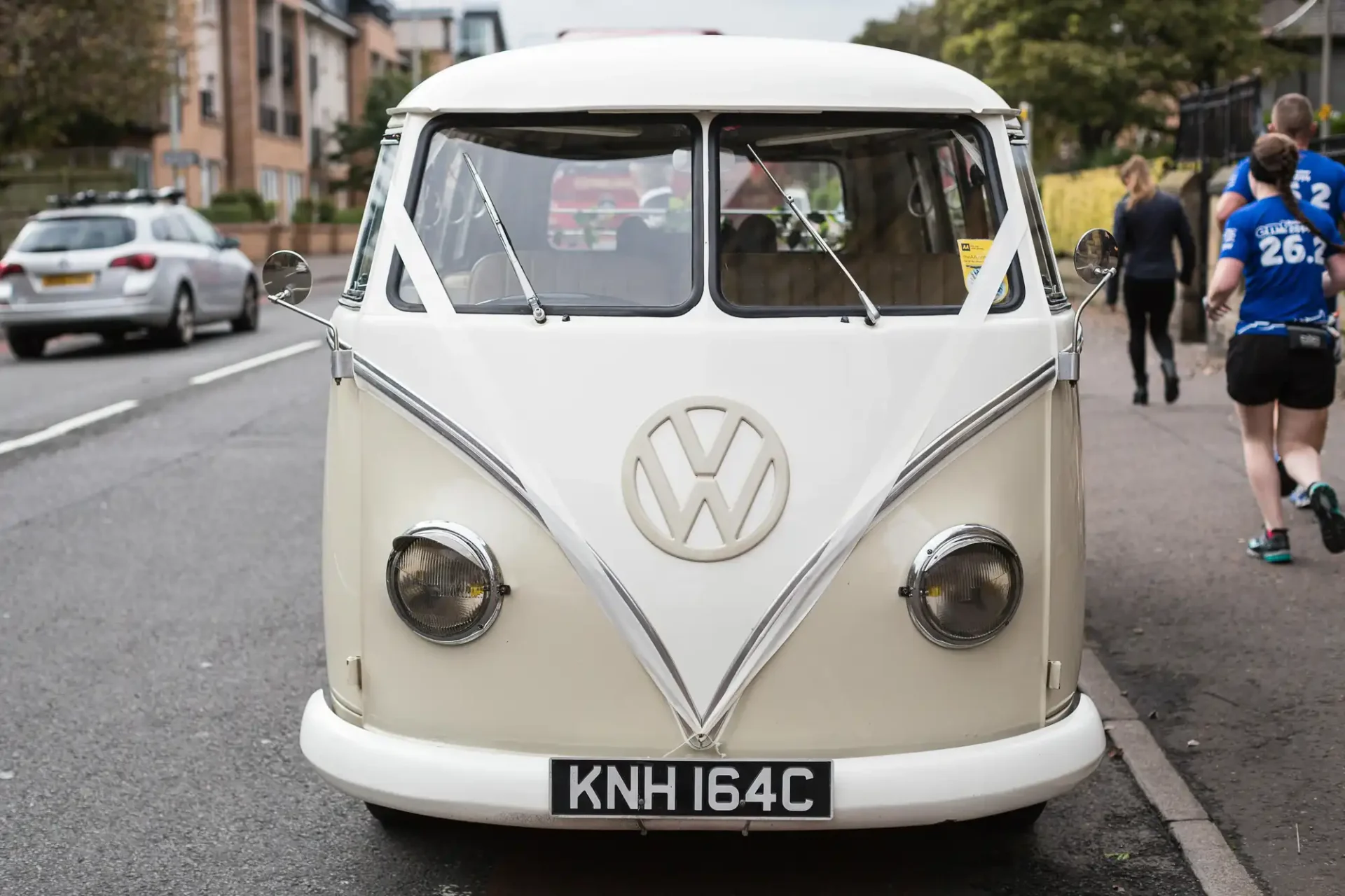 Front view of a vintage white volkswagen van with the logo visible on the front, parked on a street with pedestrians in the background.