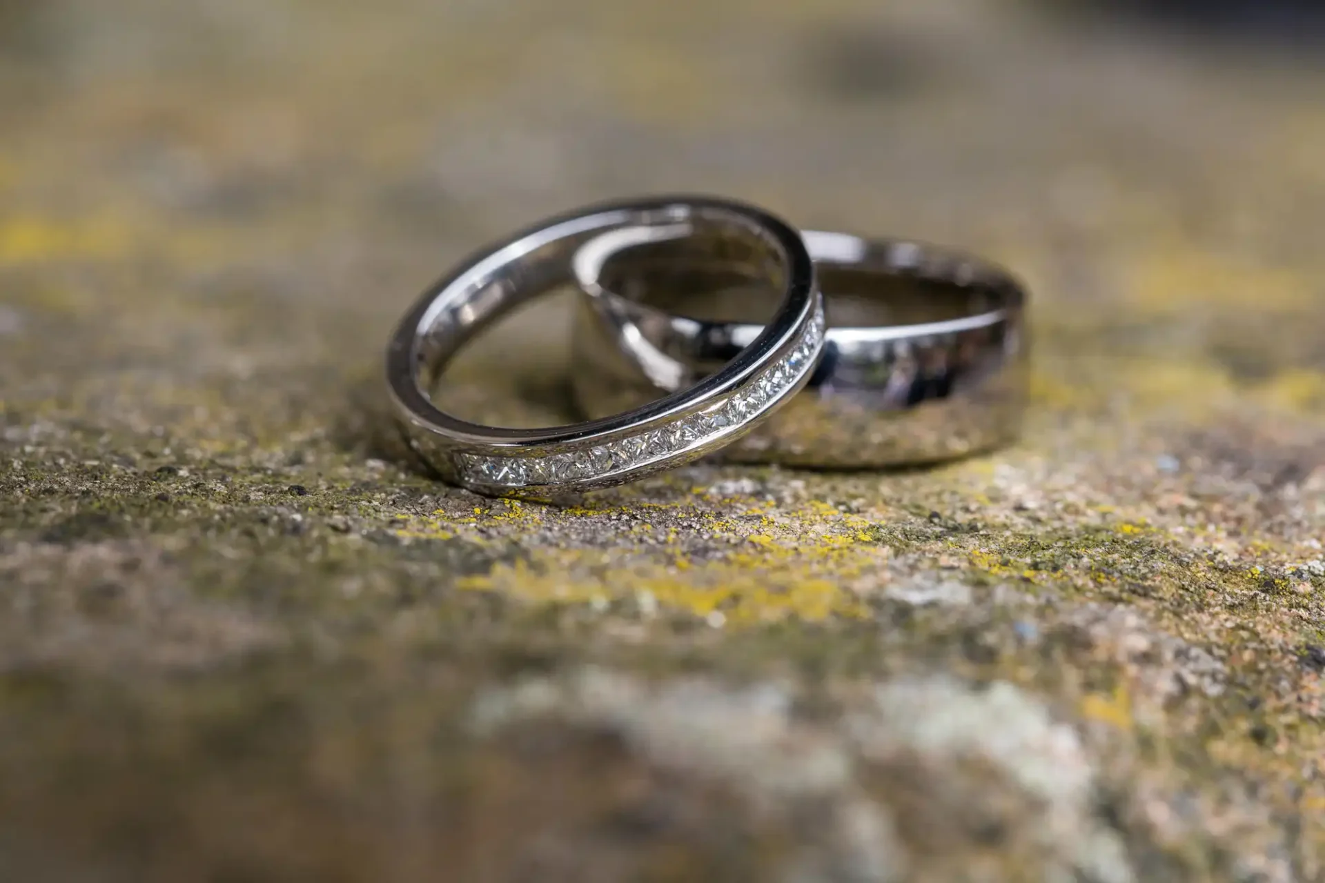Two wedding rings on a rough, mossy surface with selective focus highlighting details on the closer ring.