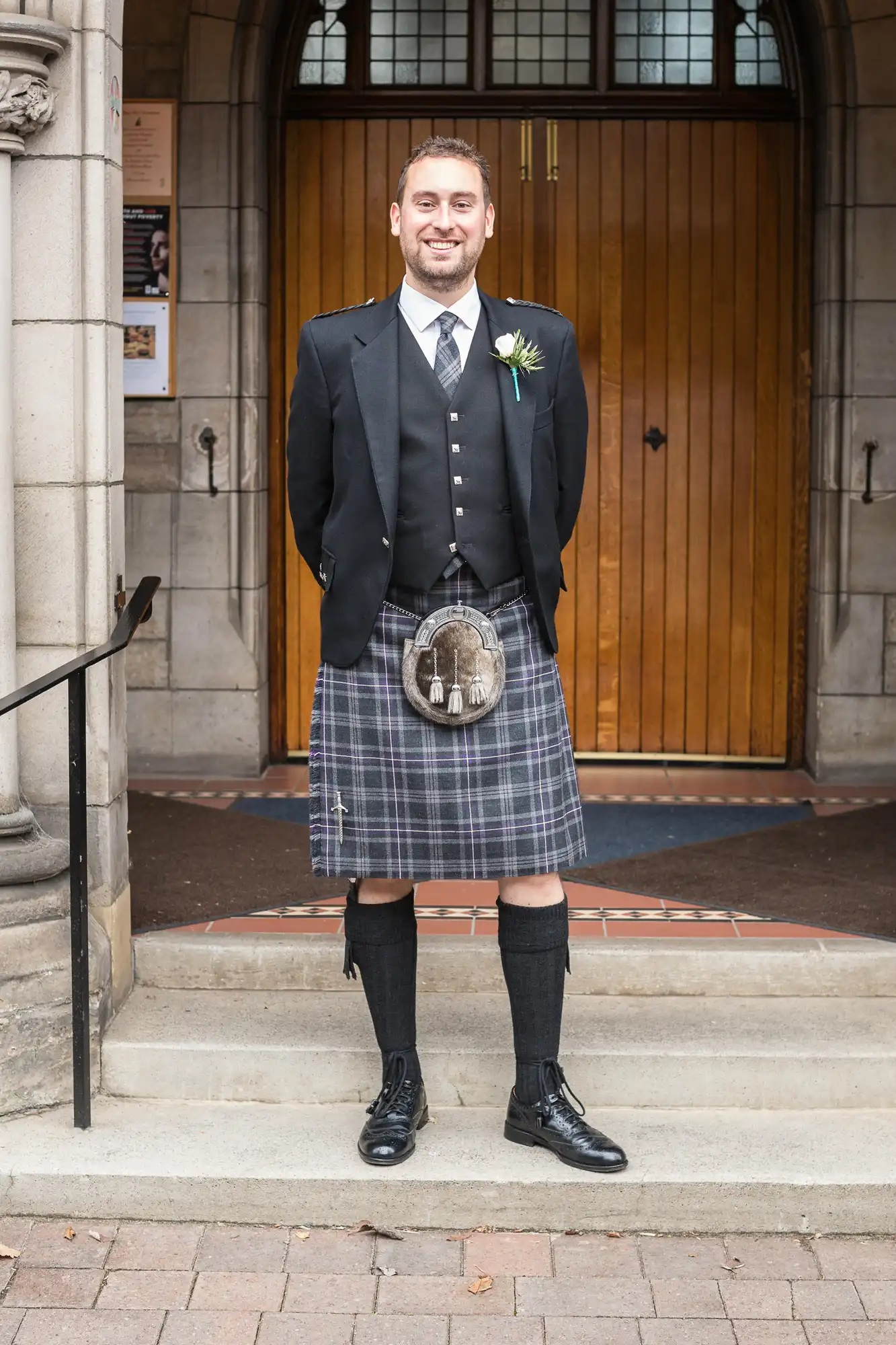 Man in traditional scottish attire smiling, standing in front of a building entrance. he wears a kilt, sporran, and a jacket with a boutonniere.