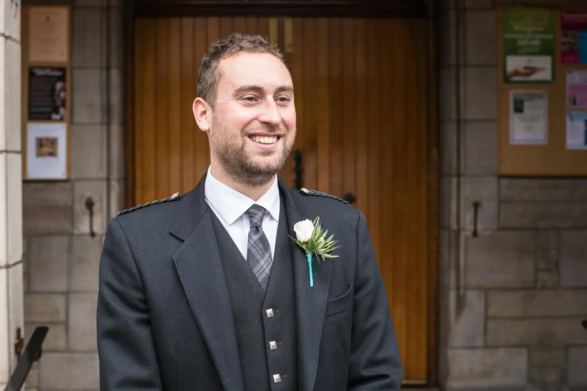 Man in a formal uniform with a boutonniere smiling outside a building with wooden doors.