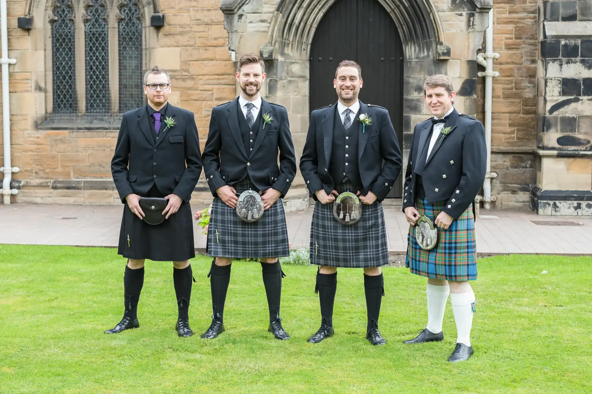 Four men in traditional scottish kilts standing in front of a stone church, smiling at the camera.