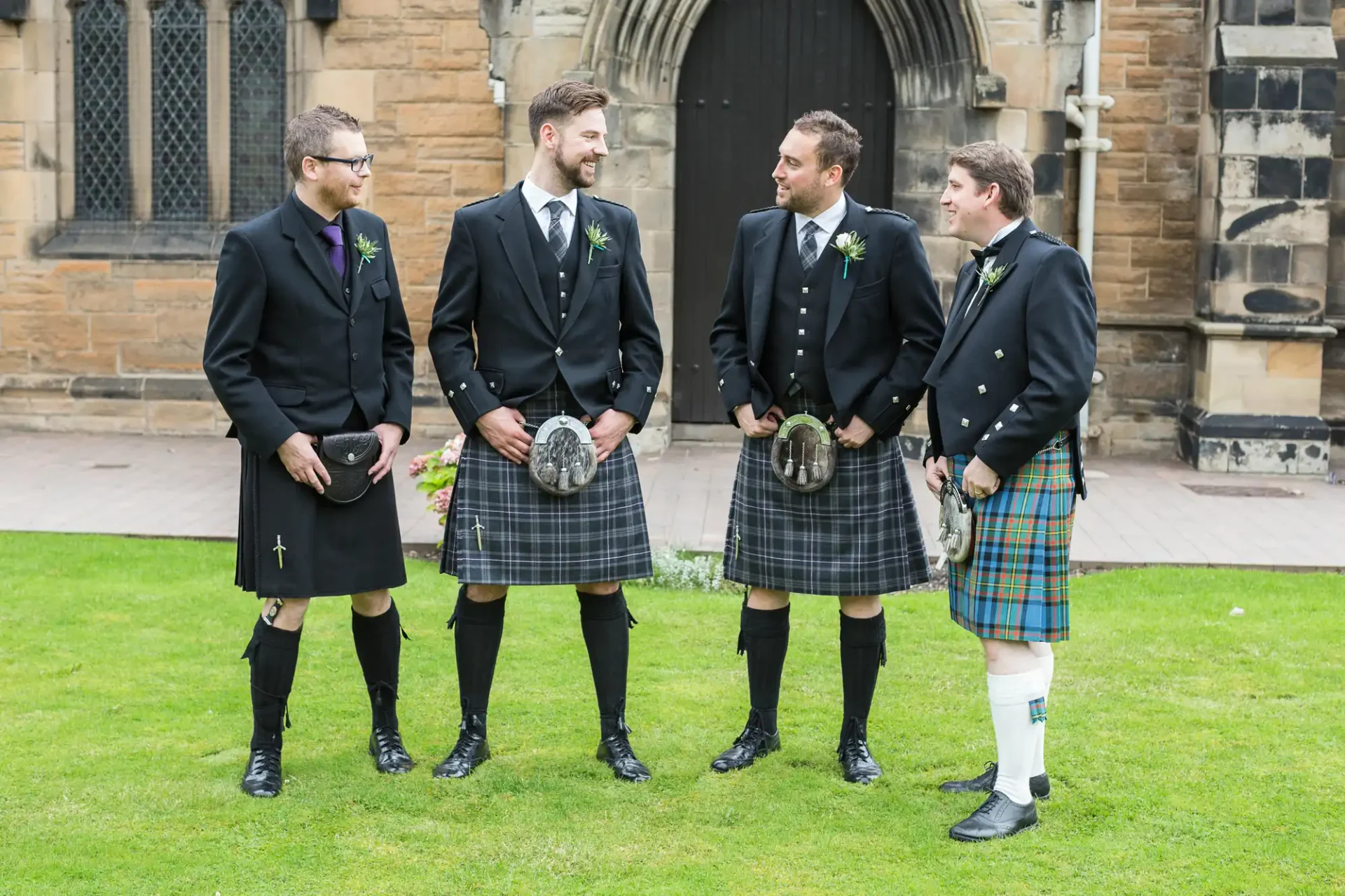 Four men in kilts and jackets, smiling and chatting outside a church, each holding a sporran.
