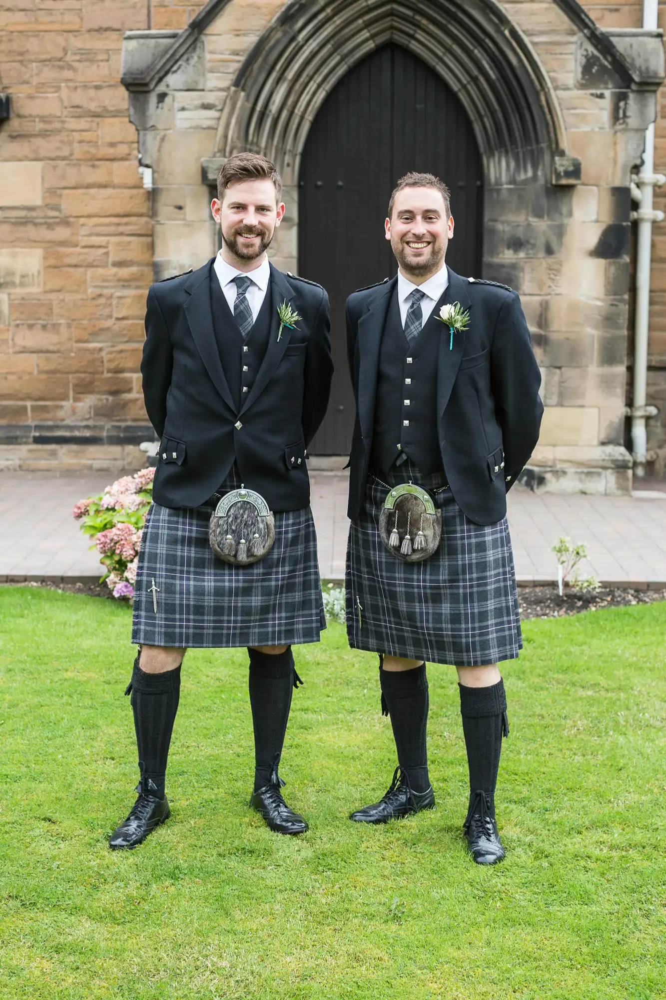 Two men in traditional scottish kilts and formal jackets smiling in front of a church entrance.