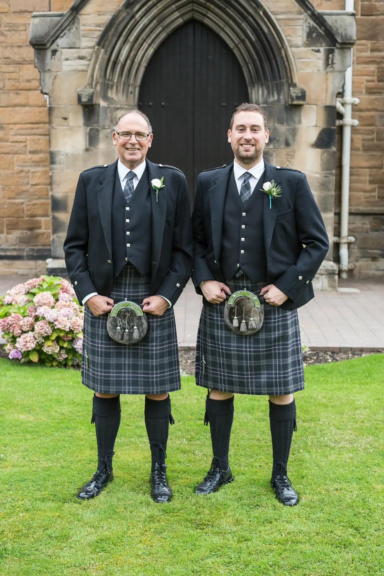 Two men in traditional scottish kilts and jackets, smiling, standing side by side in front of a stone church with flowers in the background.
