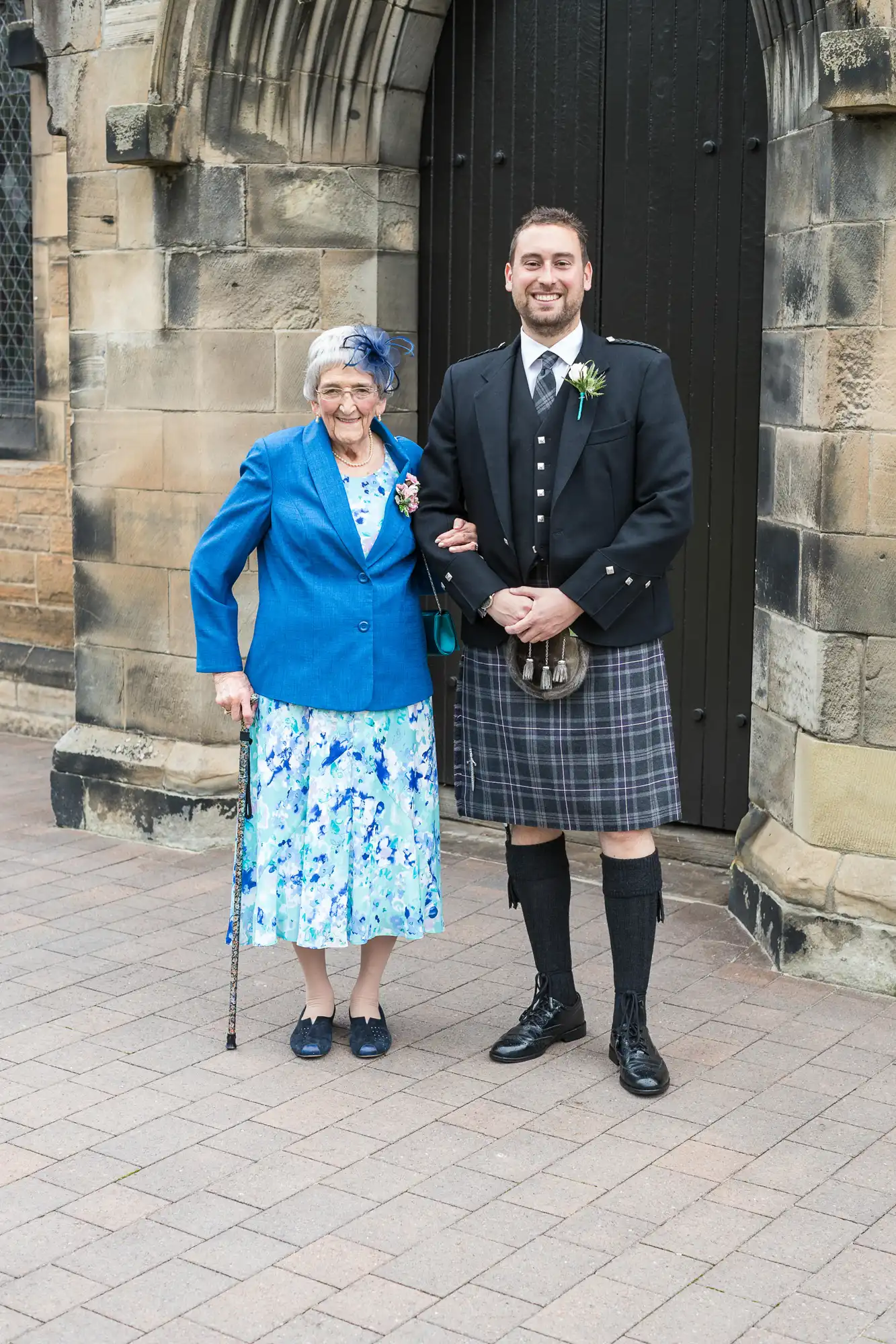 Elderly woman in blue jacket and floral dress with a younger man in traditional scottish kilt, both smiling outside a stone building.